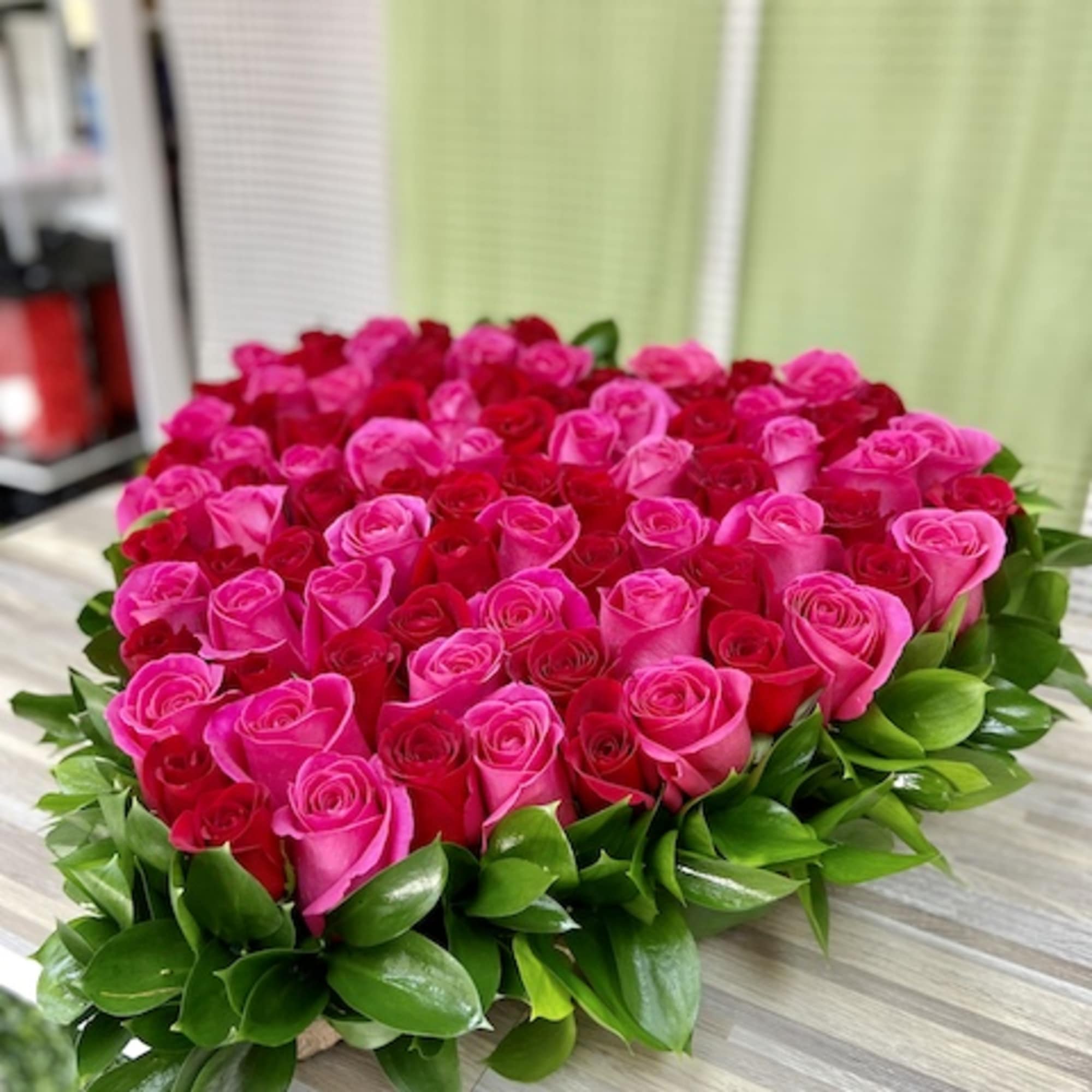 Heart-shaped arrangement of pink and red roses edged with glossy green foliage on a wooden surface