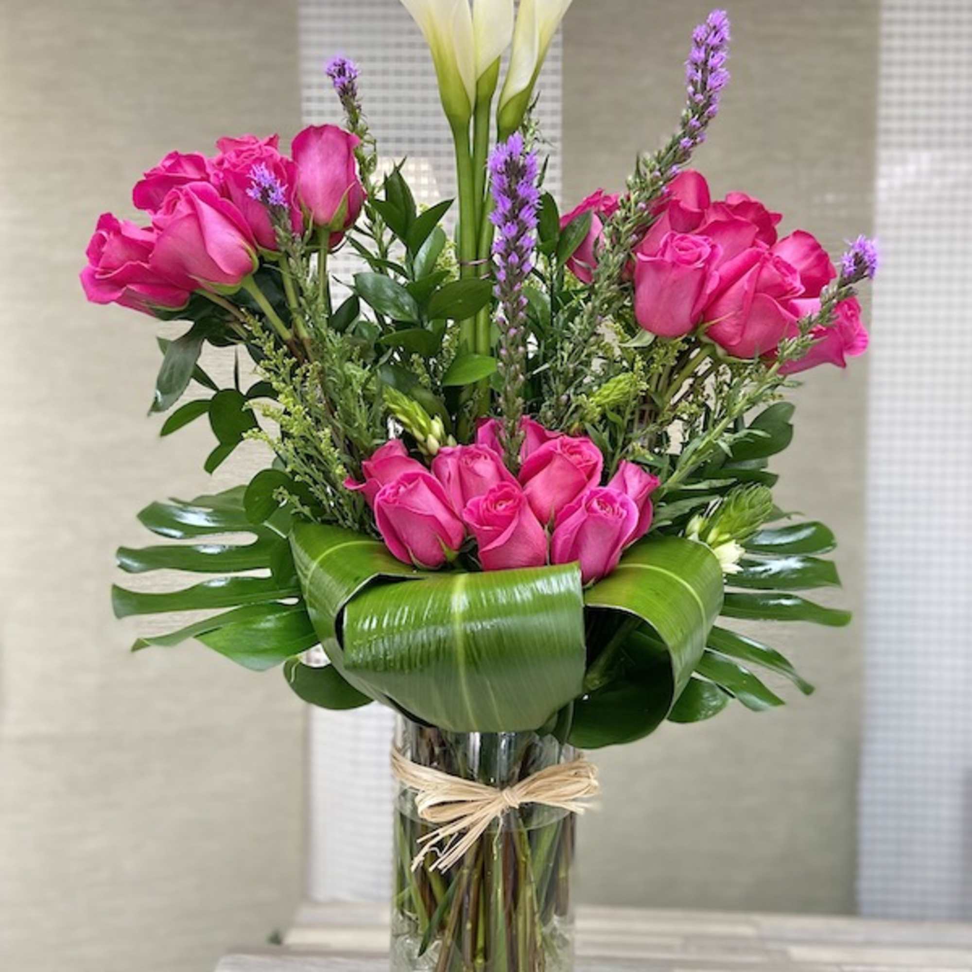 Tall arrangement of pink roses, white calla lilies, and purple flowers in a glass vase with large folded leaves