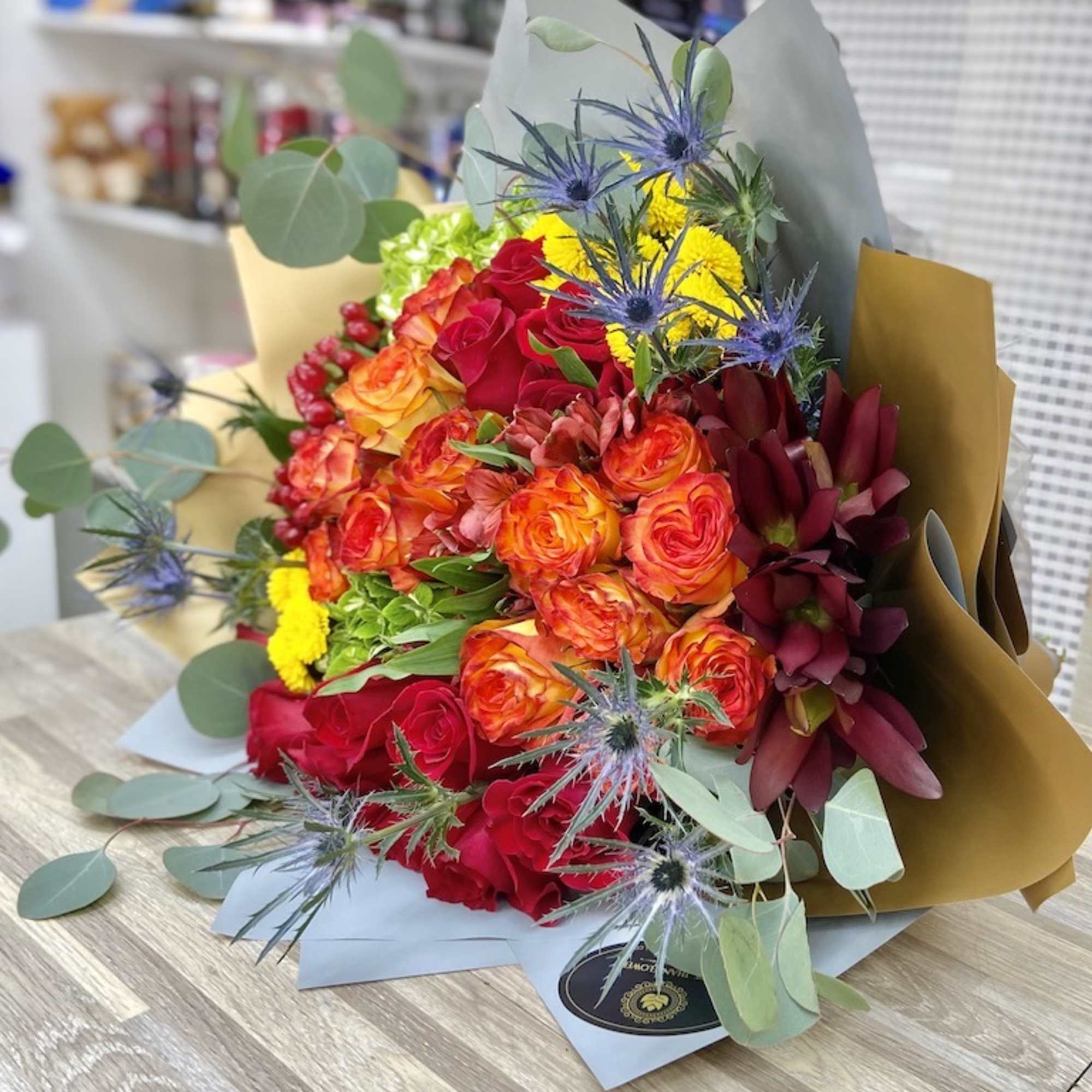 Large hand-tied bouquet of red and orange roses with yellow blooms and blue thistles in kraft paper wrap
