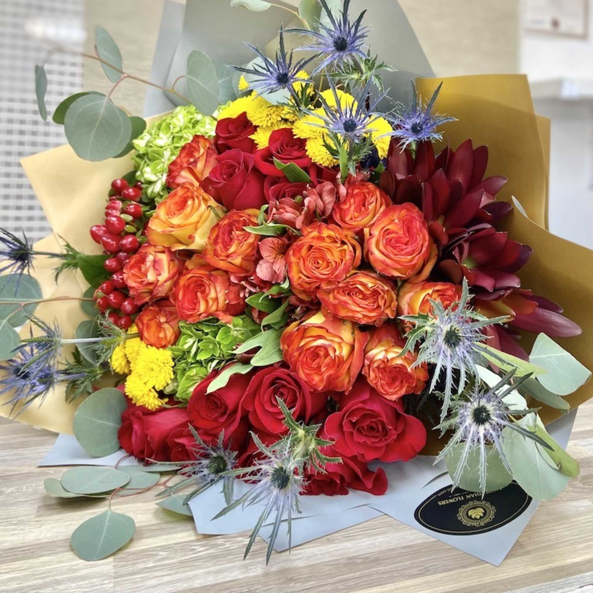 Hand-tied bouquet of red and orange roses with yellow blooms and blue thistles in kraft paper wrap
