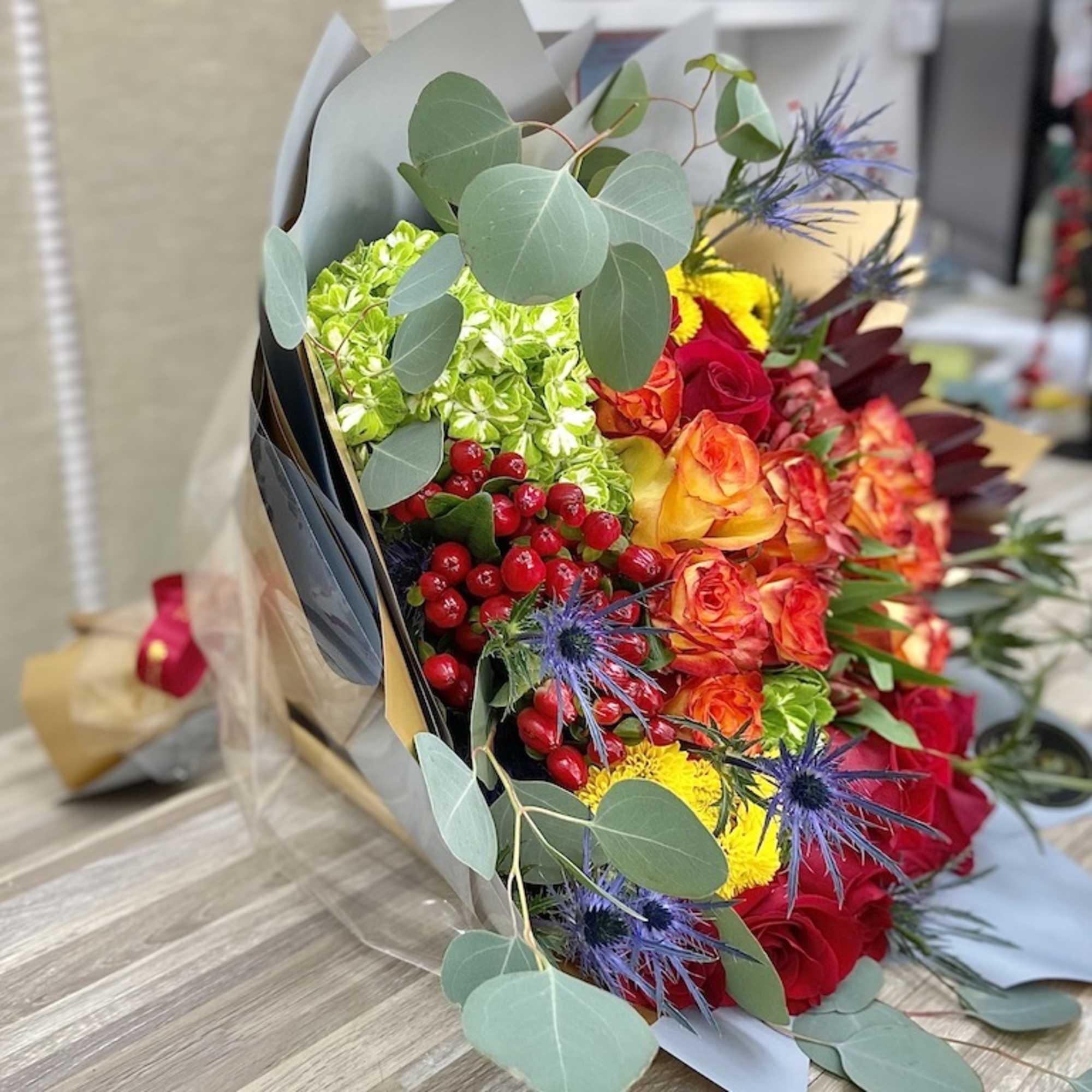 Wrapped bouquet of red and orange roses with yellow blooms, green hydrangea, berries, and blue thistles