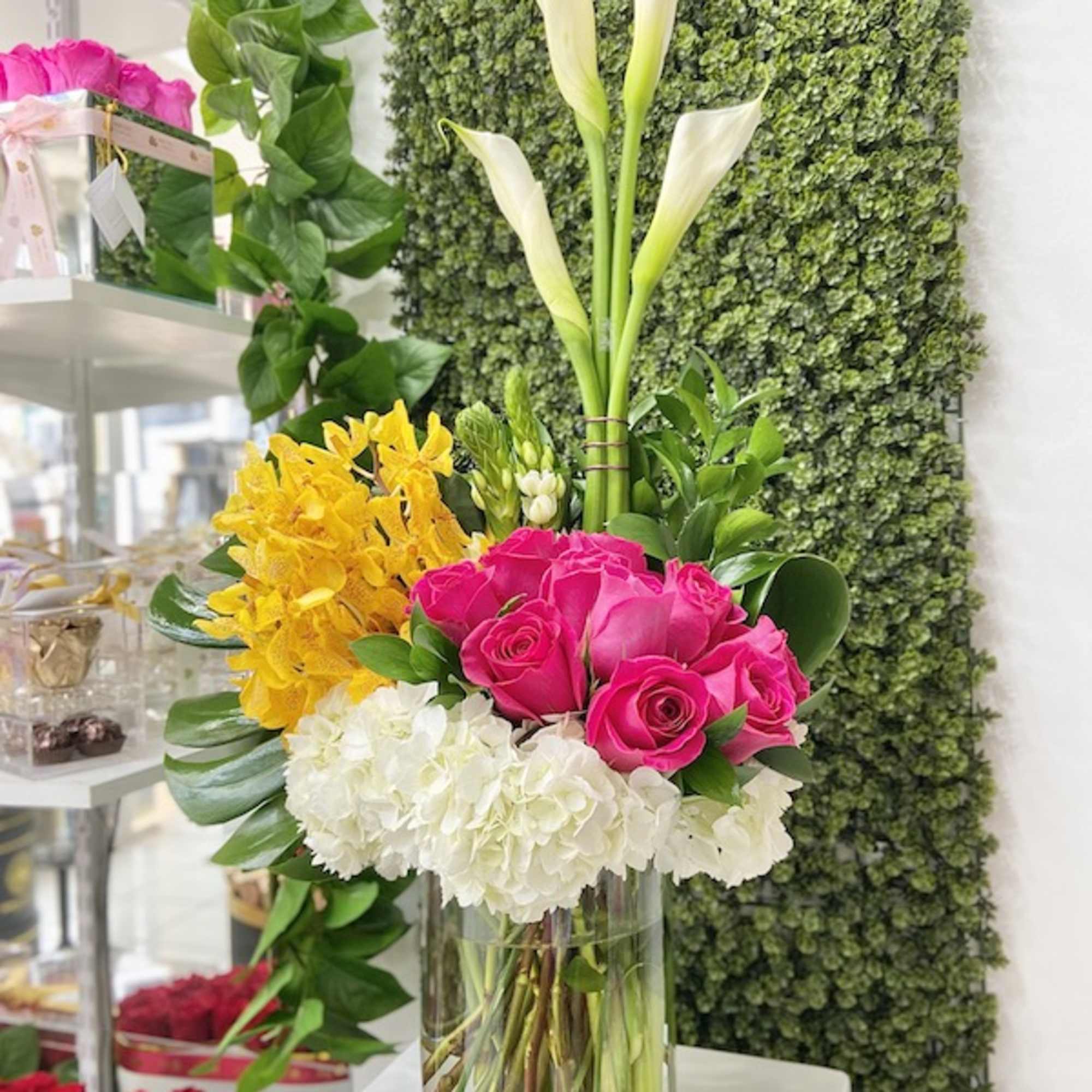 Tall mixed arrangement with white calla lilies, hot pink roses, yellow orchids, and white hydrangeas in a glass vase