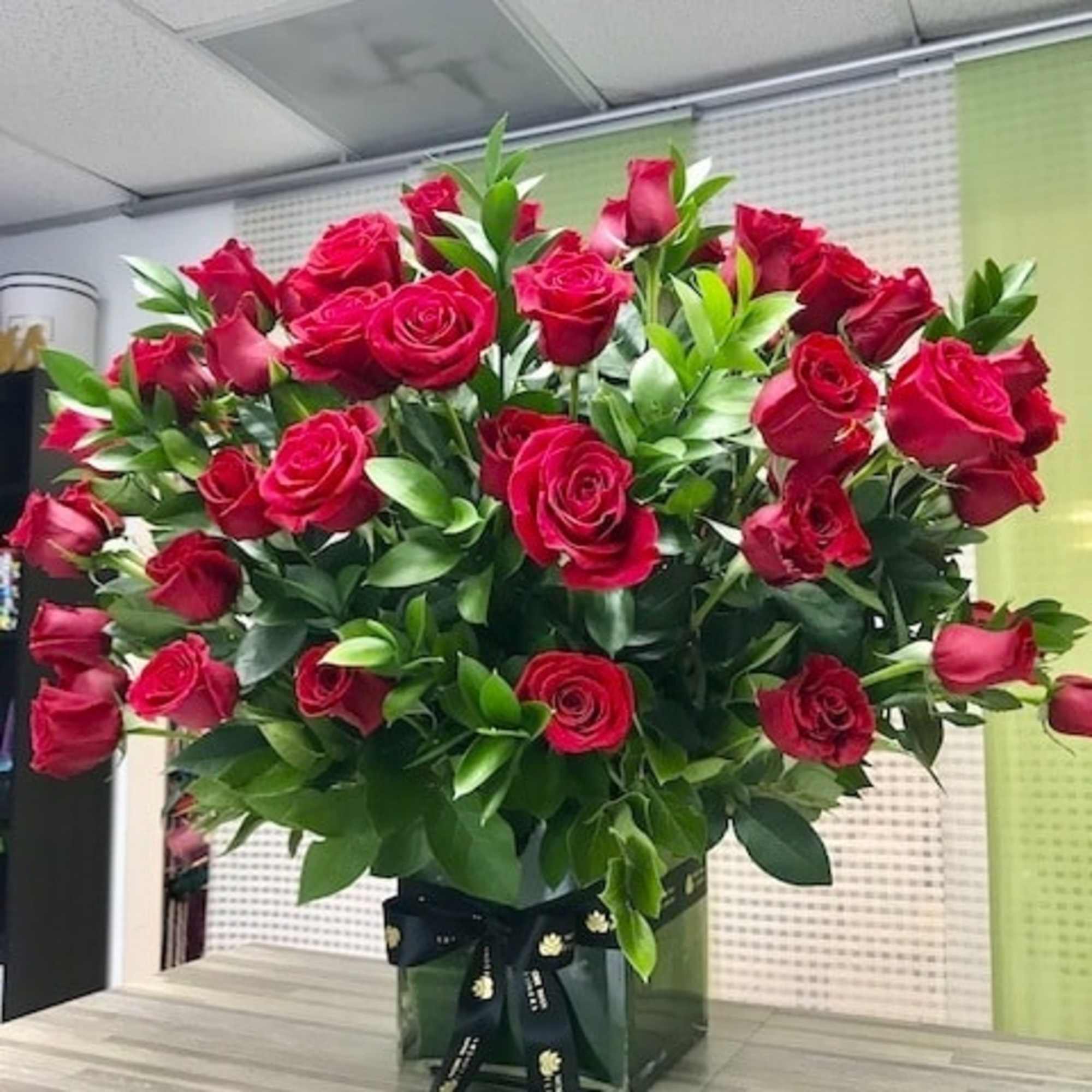 Large arrangement of red roses with abundant greenery in a clear glass cube vase tied with a black ribbon.