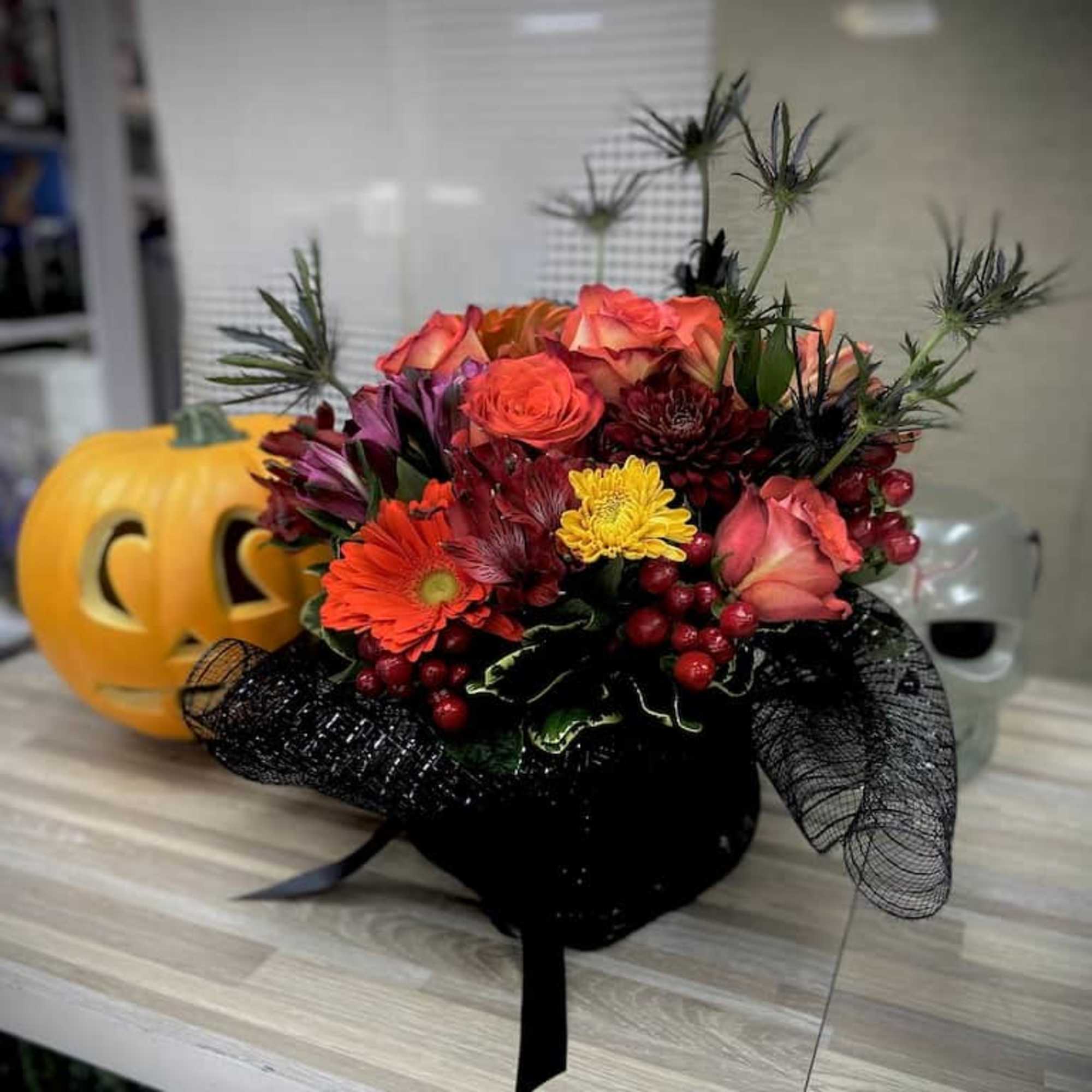 Halloween-themed orange and red floral arrangement with berries in a black container beside a jack-o-lantern.