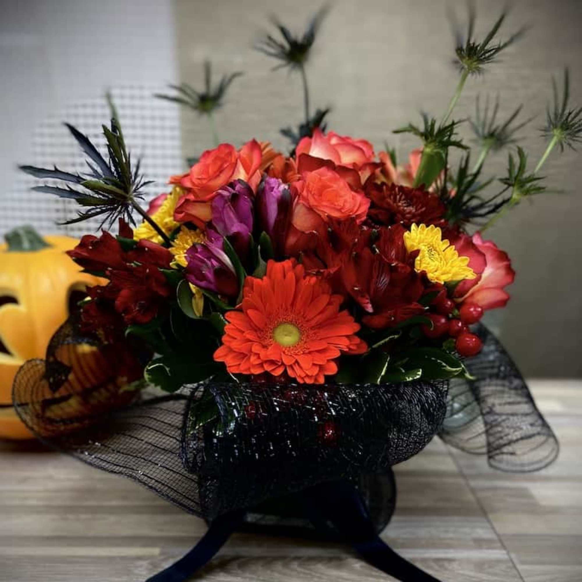 Low arrangement of orange gerbera, red and yellow blooms in a glass vase with black mesh wrap