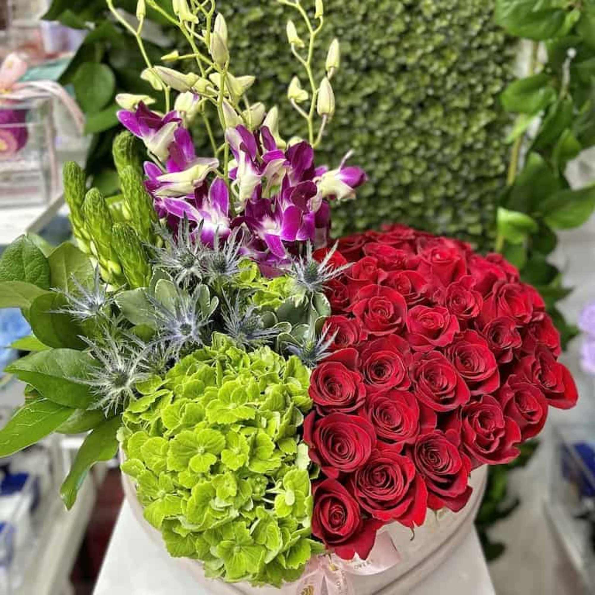 Heart box of red roses, green hydrangeas, purple orchids, and accent blooms with a pink ribbon