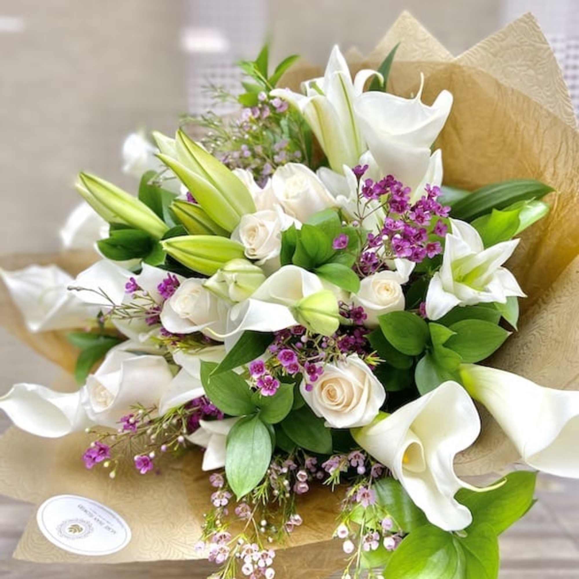 Hand-tied bouquet of white calla lilies, roses, and lilies with purple filler flowers in kraft paper wrap