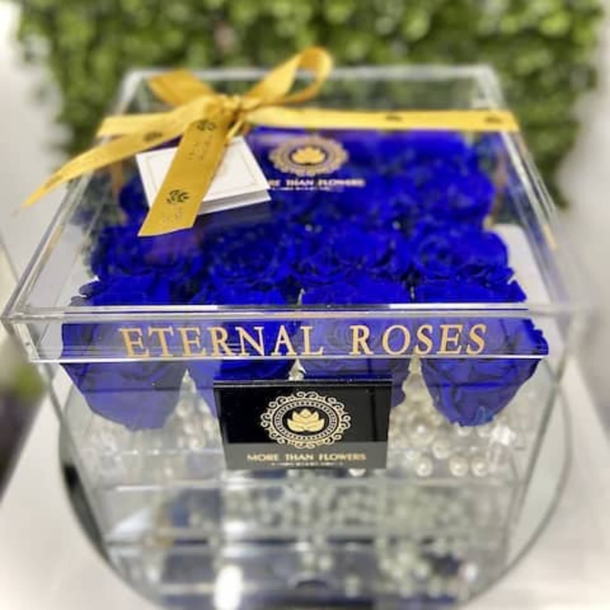 Clear acrylic box of vivid blue roses labeled Eternal Roses with a gold ribbon on top