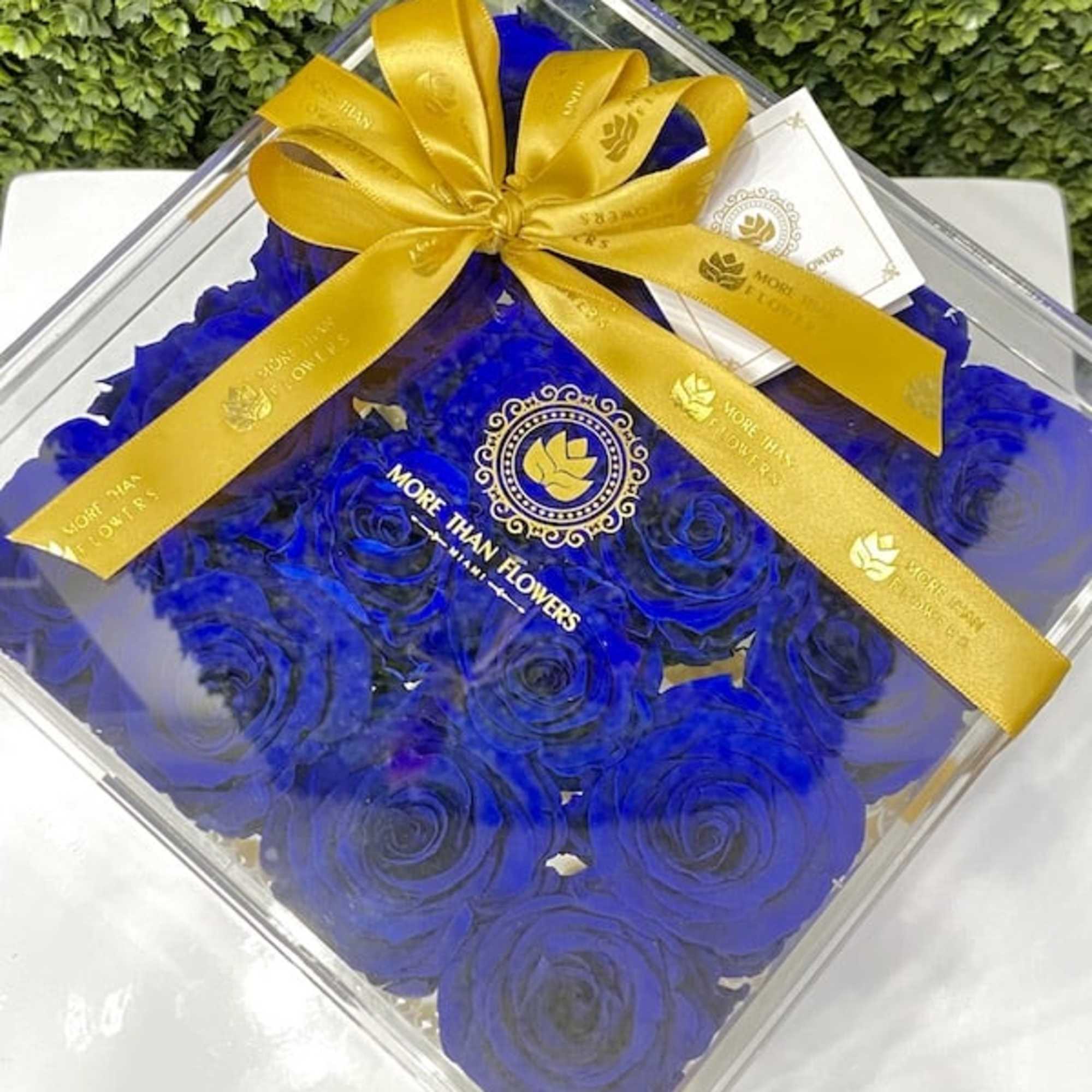 Square clear box of vivid blue roses topped with a yellow ribbon and small card.