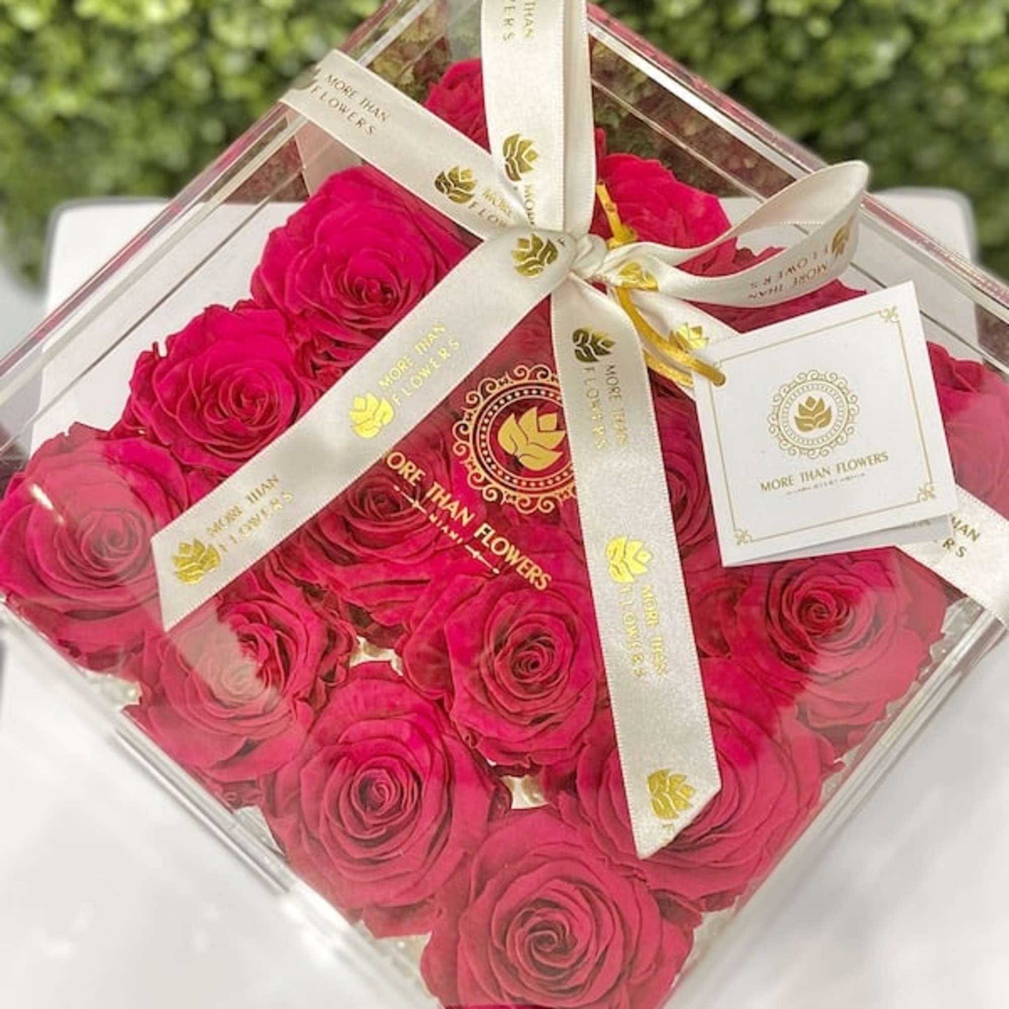 Bright pink roses arranged in a clear acrylic box tied with a cream ribbon and tag