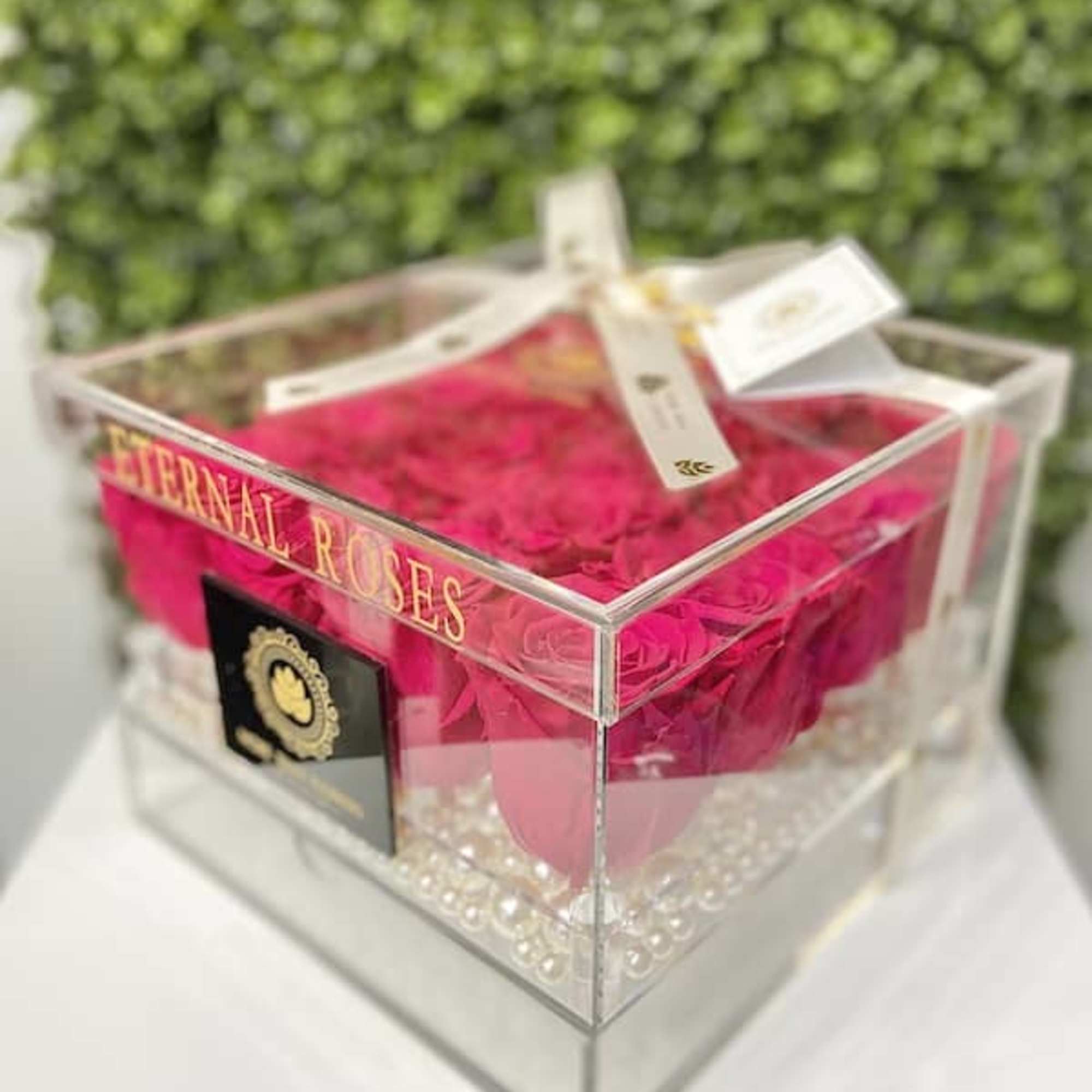 Clear acrylic box filled with bright pink roses and white pearls, tied with a ribbon.