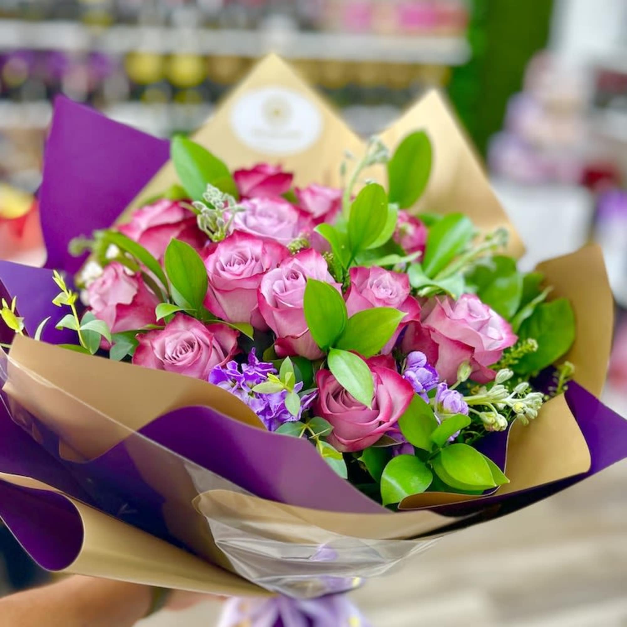 Handheld bouquet of pink roses with purple accents wrapped in tan and purple paper with a ribbon