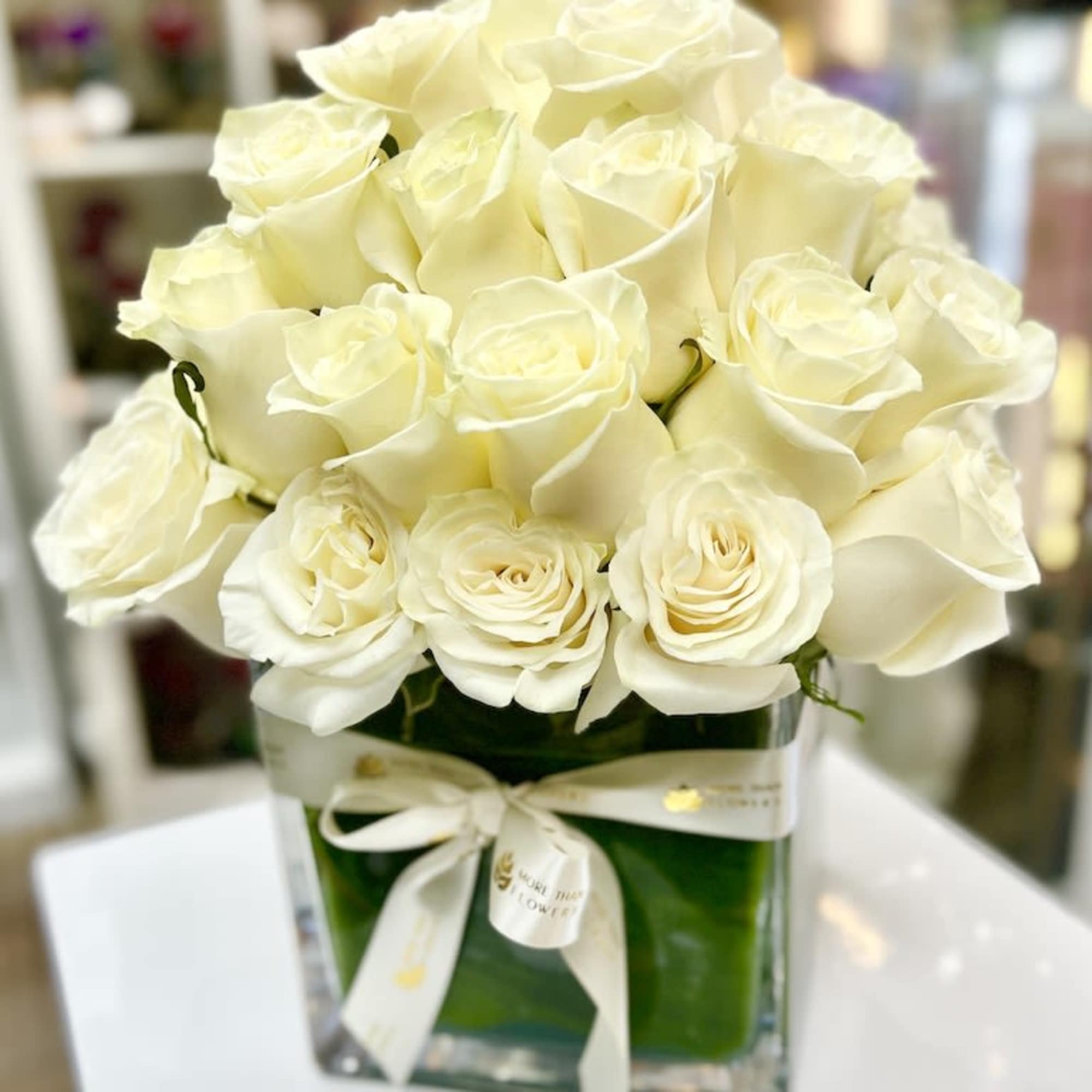 Compact arrangement of white roses in a clear glass cube vase wrapped with a cream ribbon