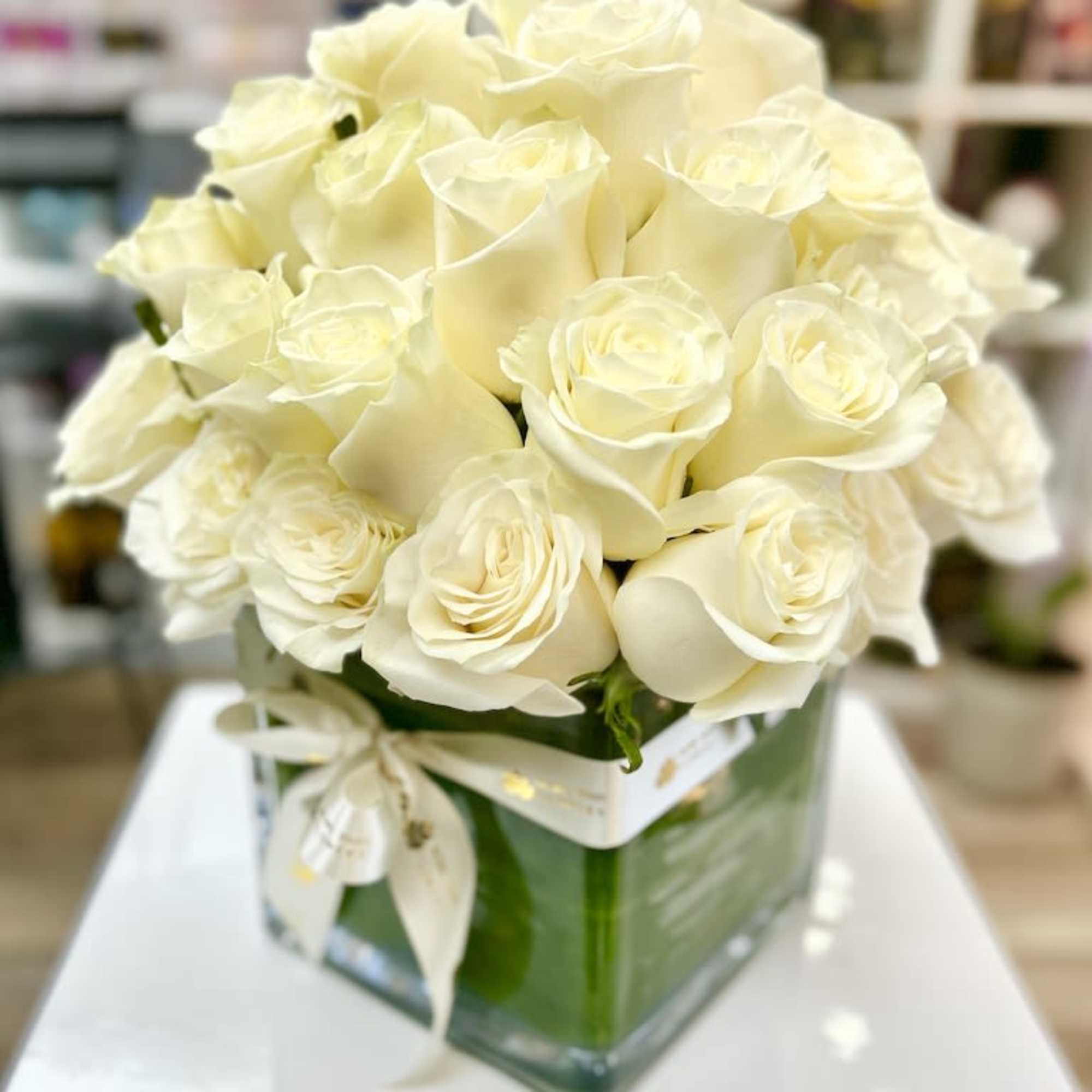 Compact arrangement of cream roses in a clear square glass vase with a ribbon bow.