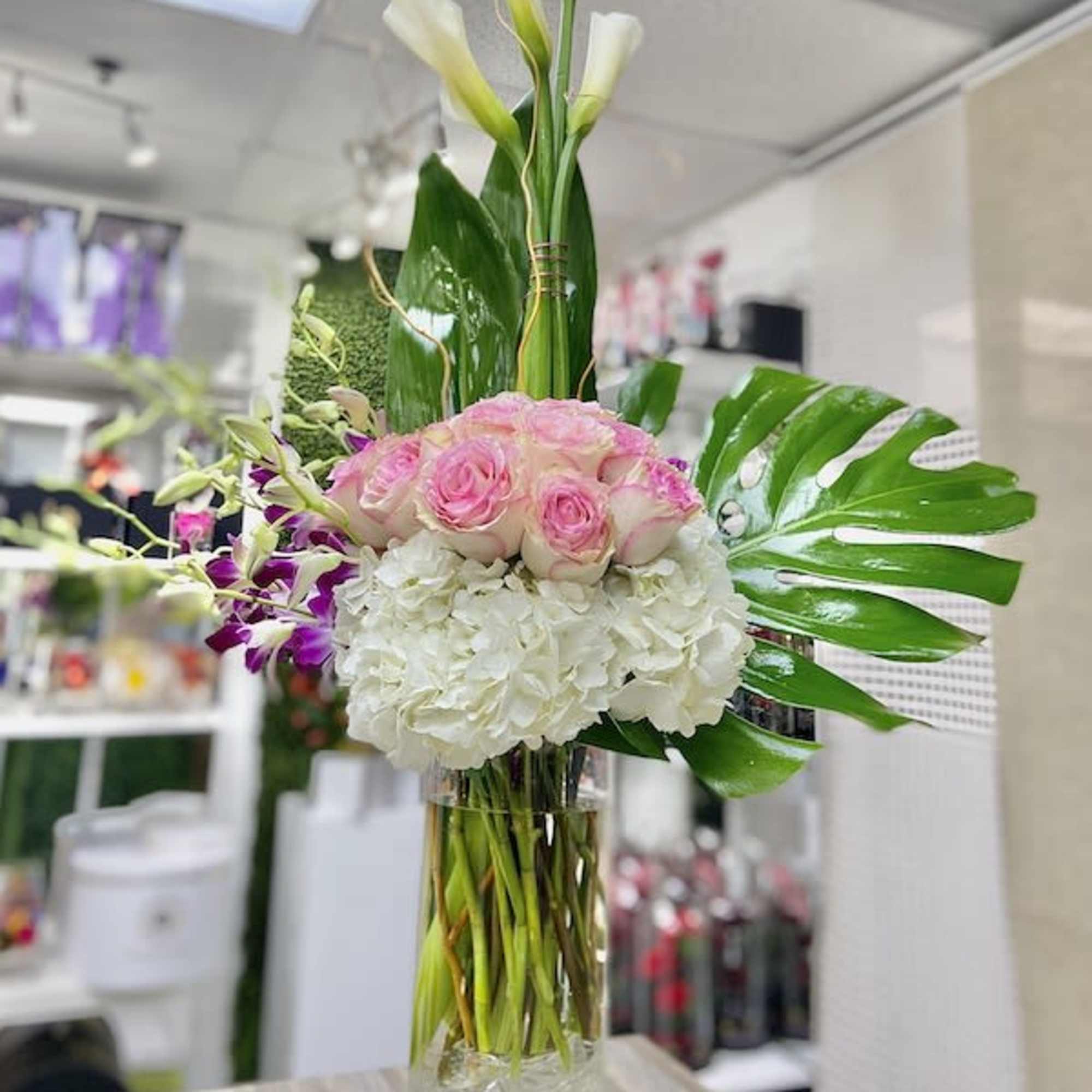 Tall arrangement of white calla lilies, pink roses, white hydrangeas and orchids in a clear glass cylinder vase.
