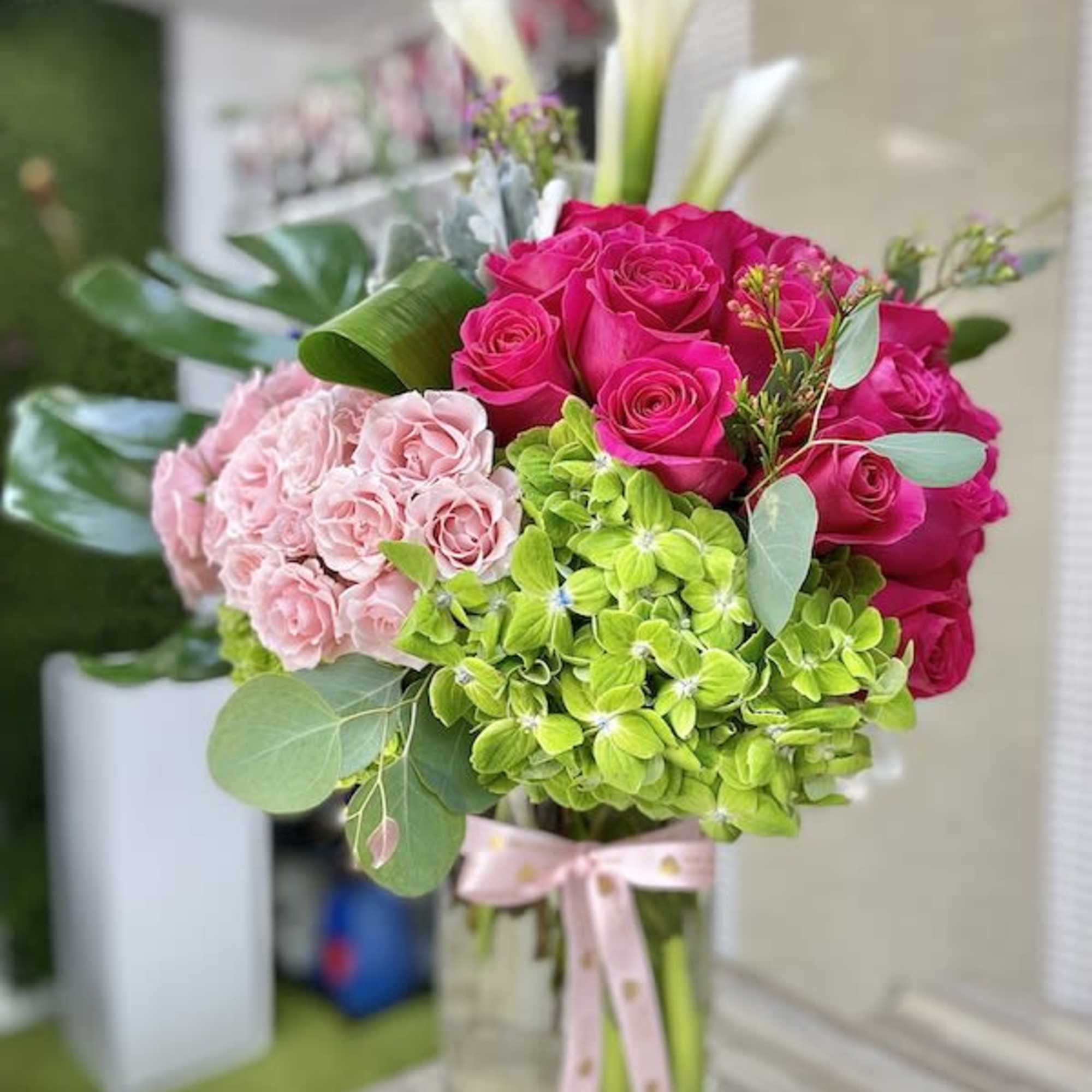 Tall glass vase arrangement with hot pink roses, pale pink blooms, green hydrangeas, and white calla lilies.