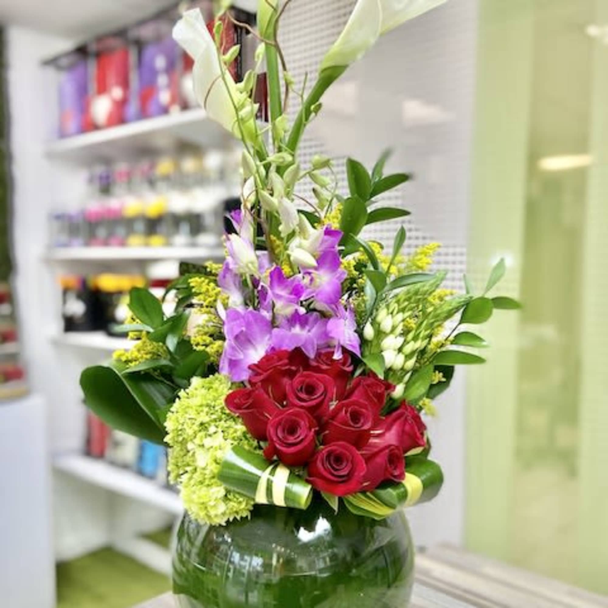 Tall arrangement of red roses, purple orchids, and white calla lilies in a round glass vase