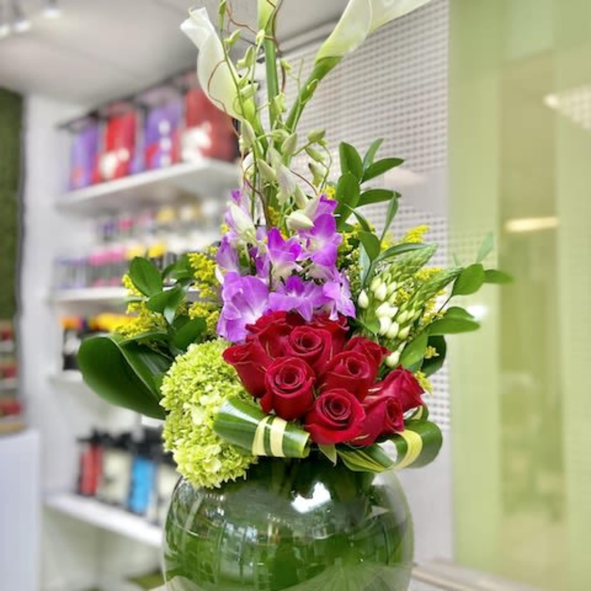 Tall mixed arrangement with white calla lilies, purple orchids, red roses, and green hydrangea in a round glass vase.