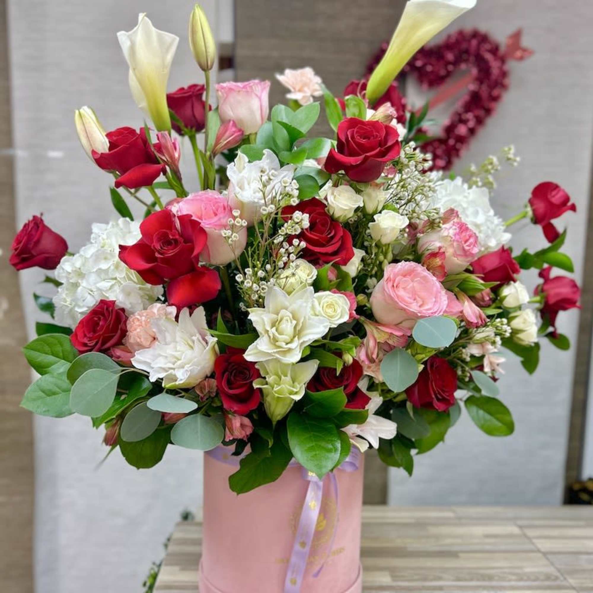Tall mixed arrangement of red and pink roses, white calla lilies and hydrangeas in a pink hatbox.