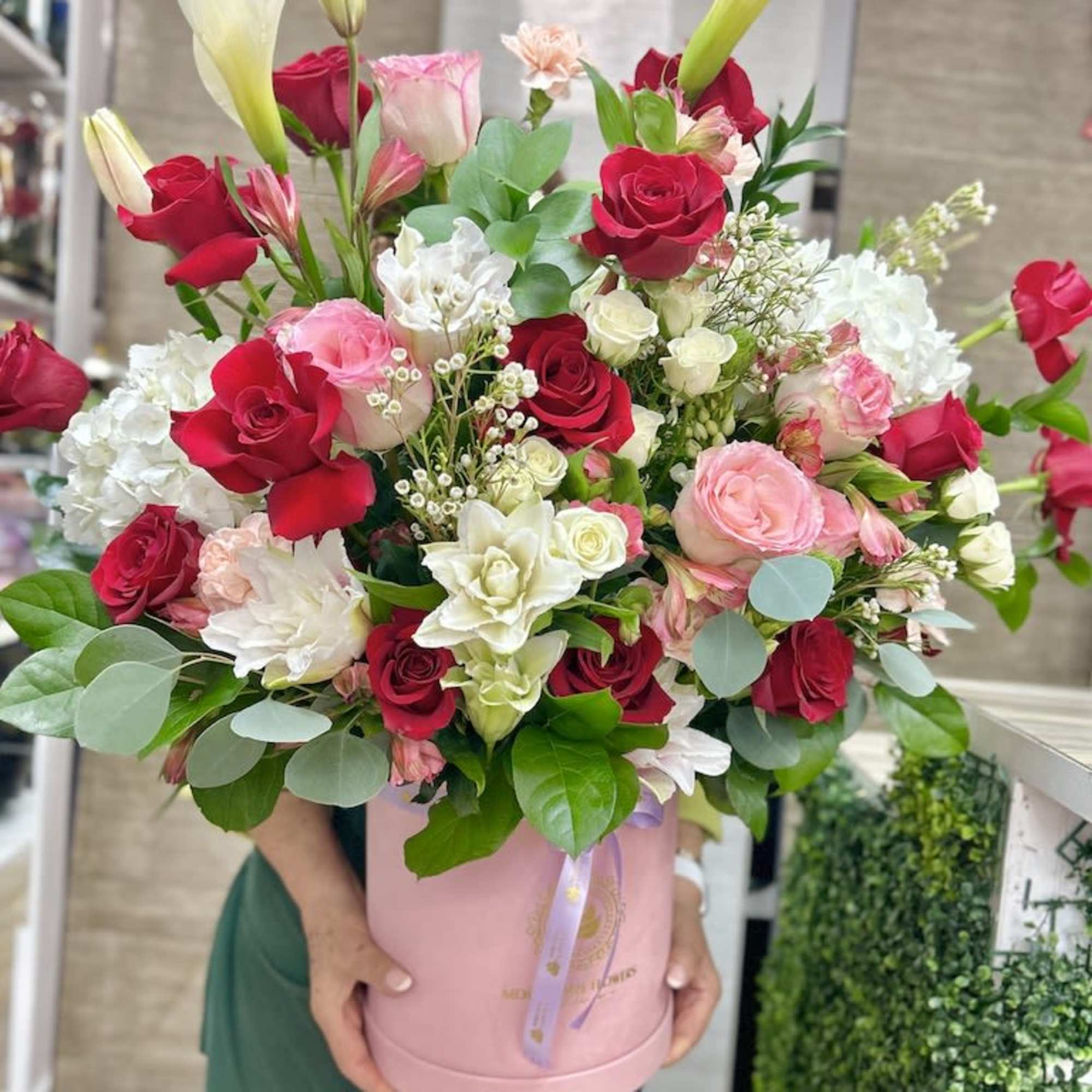 Tall arrangement of red and pink roses with white blooms and calla lilies in a pink hatbox container