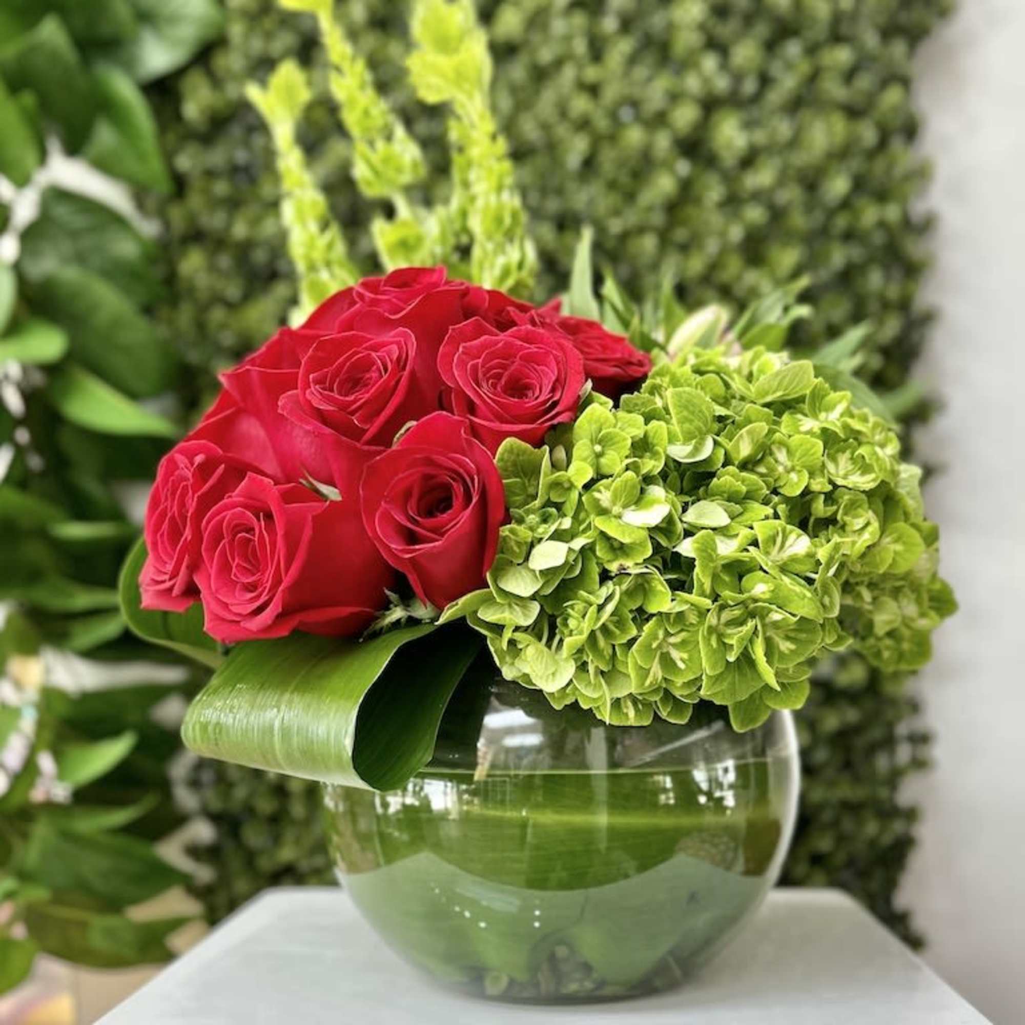 Low arrangement of red roses and green hydrangeas in a clear round vase wrapped with broad leaves.