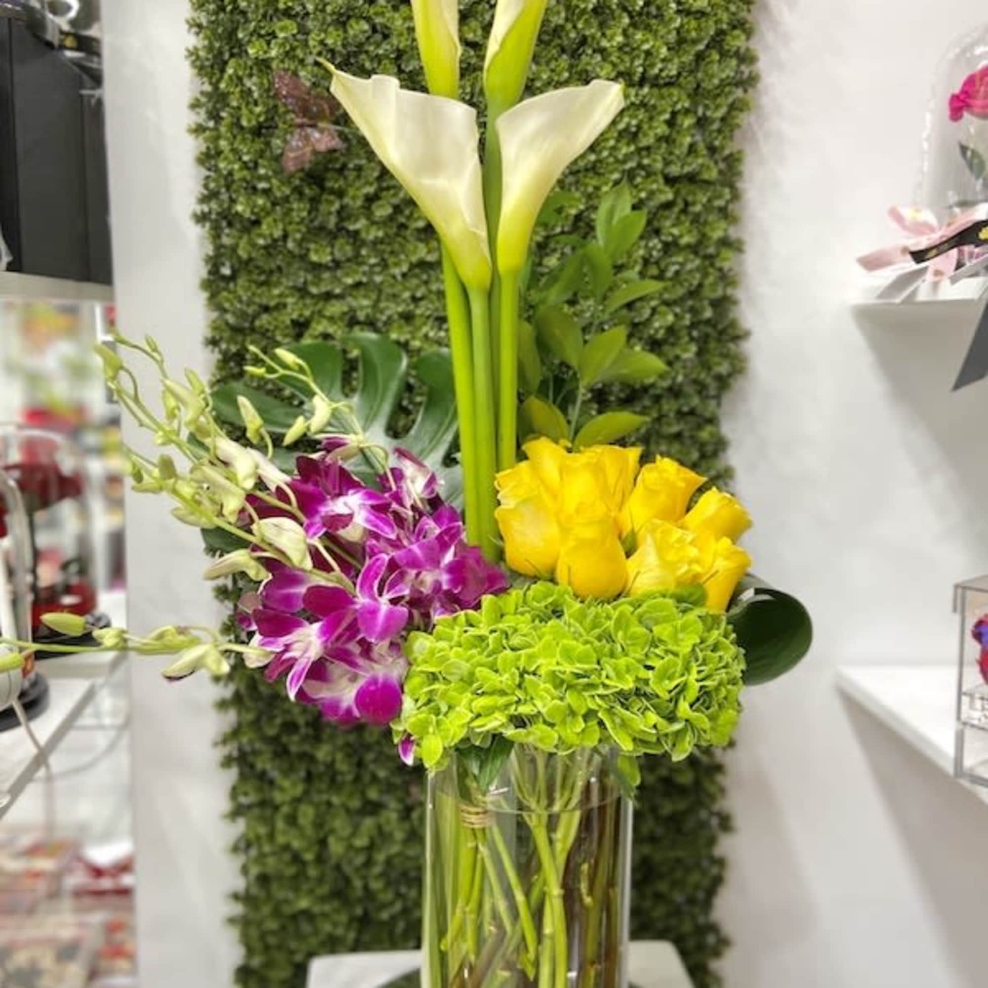 Tall arrangement of white calla lilies, green hydrangea, yellow roses, and purple orchids in a clear glass vase