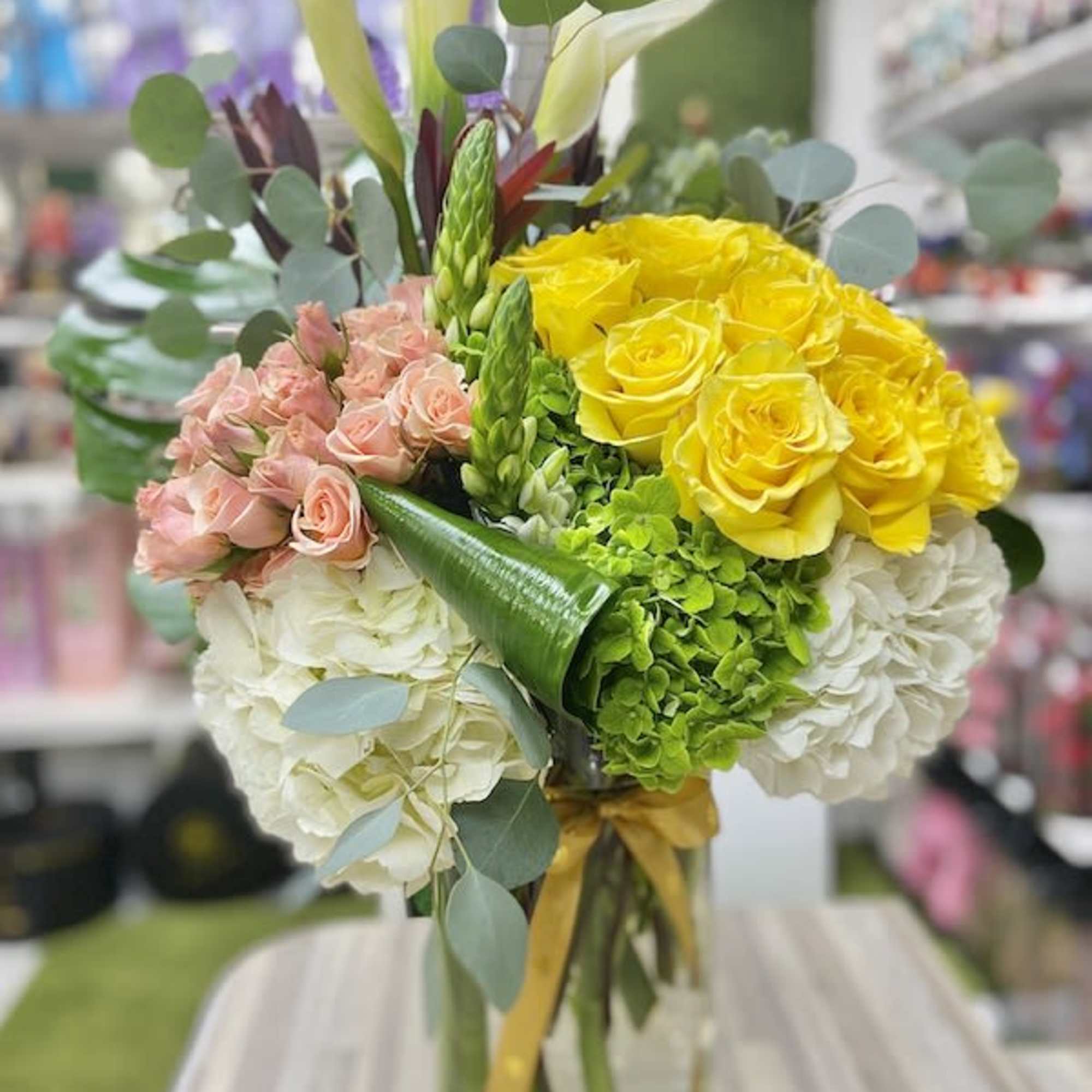 Tall mixed arrangement of yellow roses, peach spray roses, white hydrangeas and calla lilies in a glass vase.