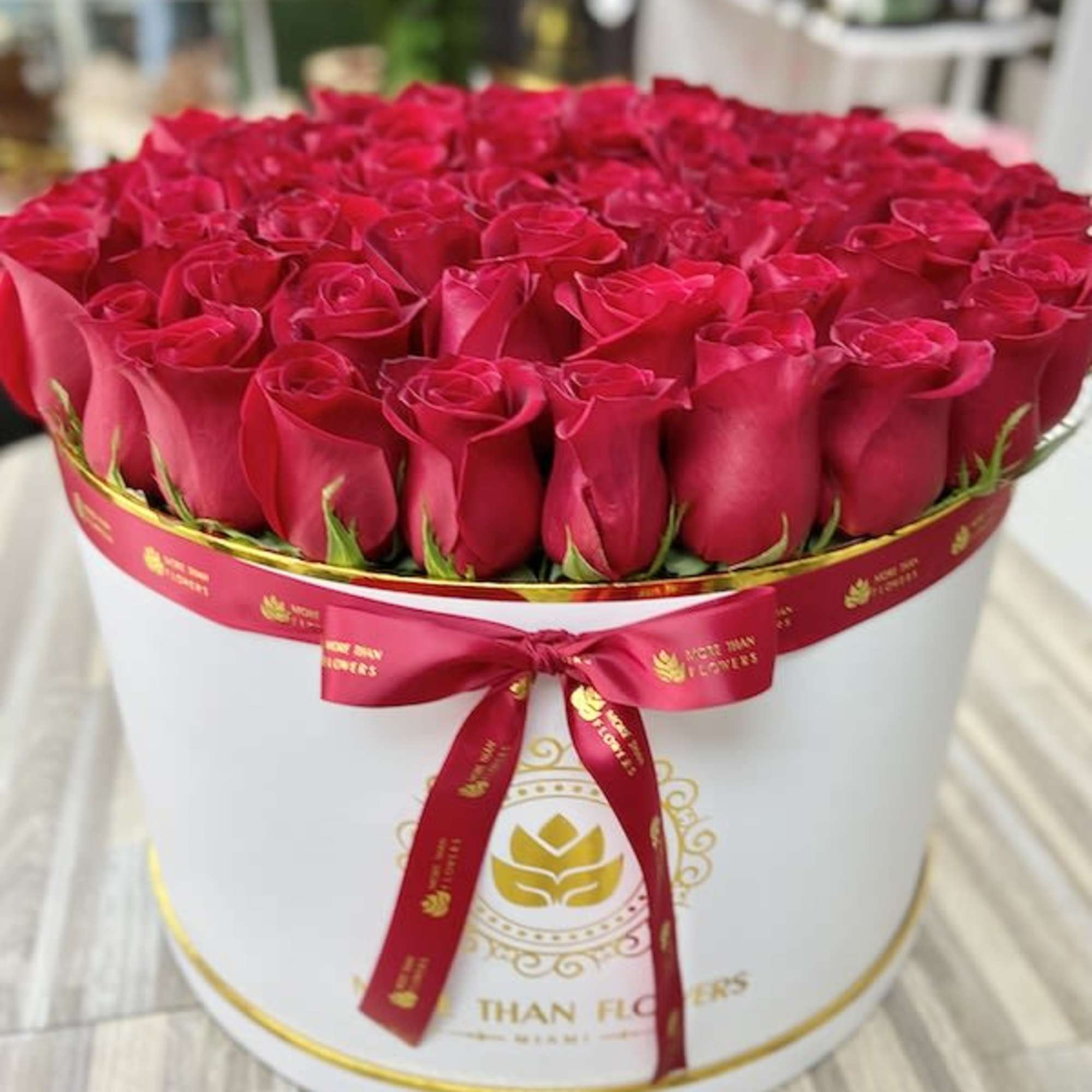Round white hatbox filled with tightly packed red roses and tied with a red ribbon.