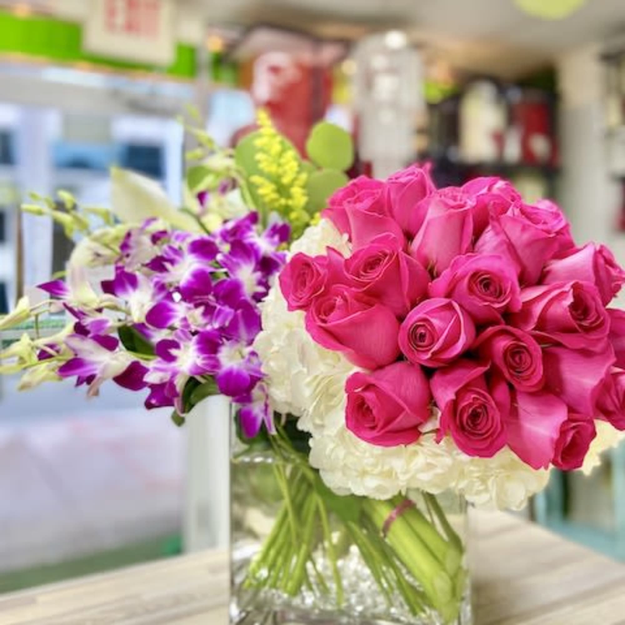 Square vase arrangement with hot pink roses, white hydrangeas, and purple orchids on a wooden surface