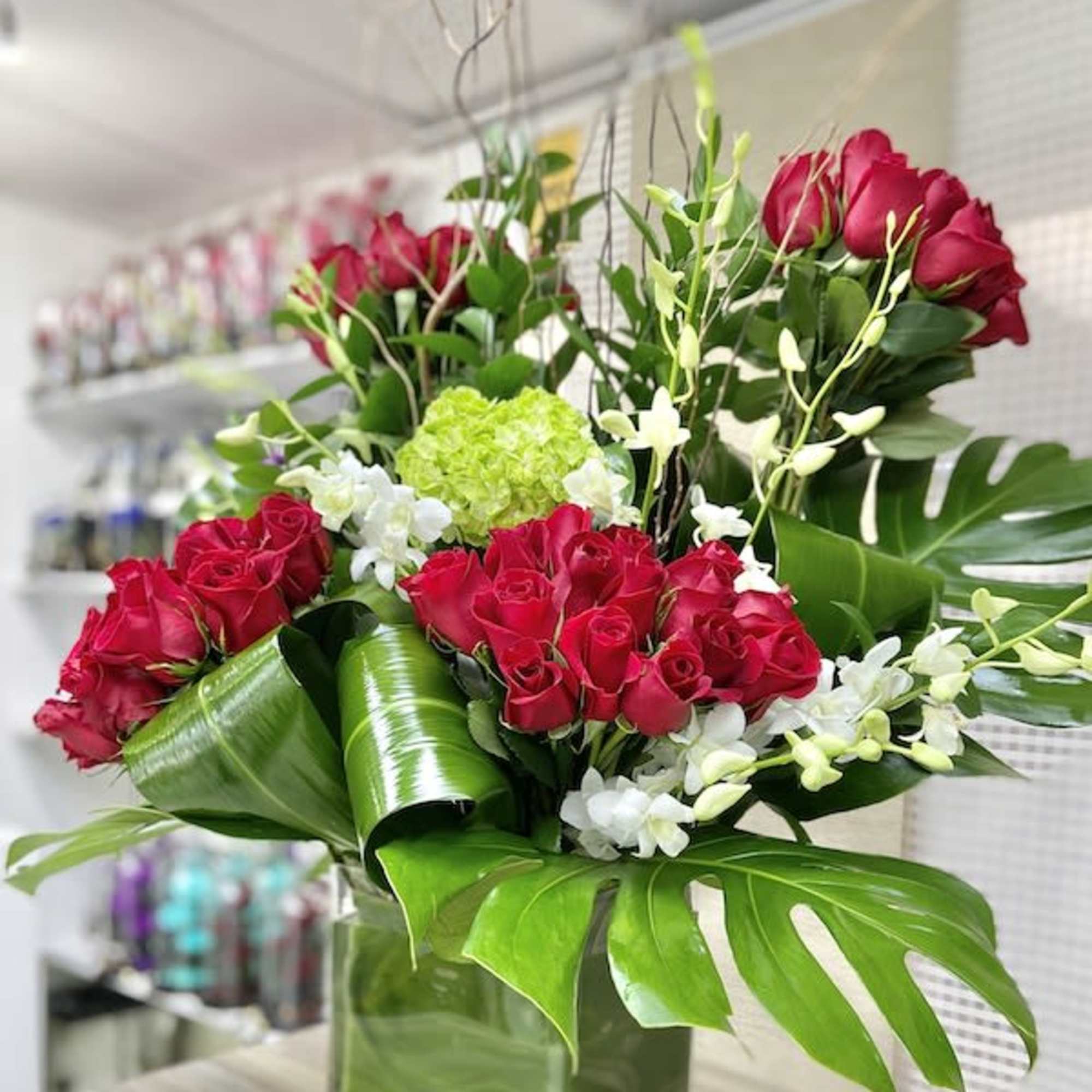 Tall arrangement of red roses, white orchids, and green hydrangea with large tropical leaves in a glass vase