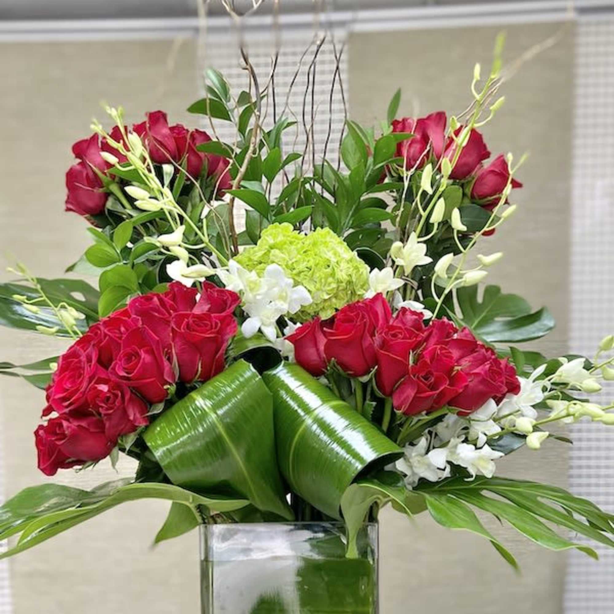 Tall modern arrangement of red roses, white orchids, and green hydrangea in a glass cube with large folded green leaves