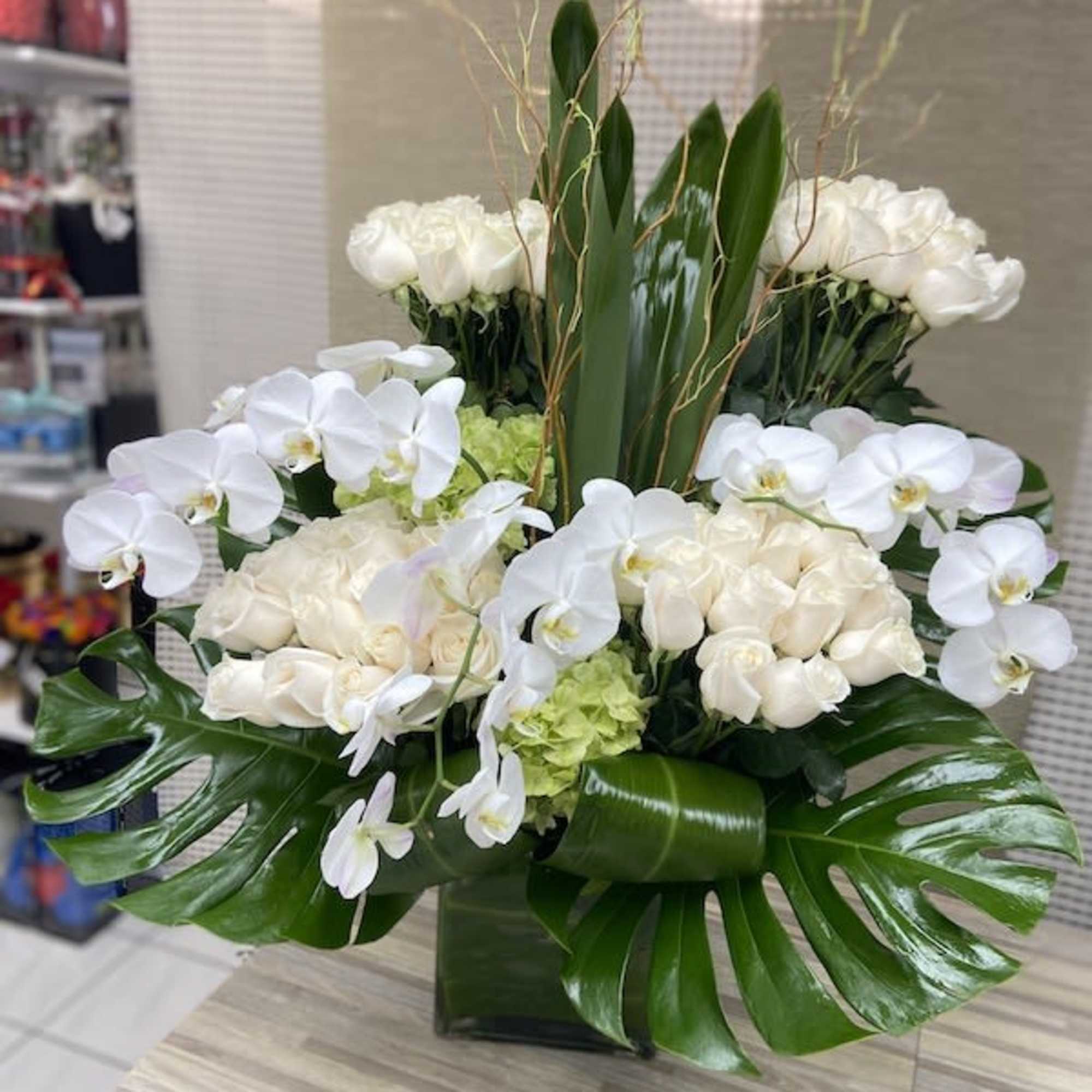 Large modern arrangement of white roses and orchids with bold tropical leaves in a square glass vase