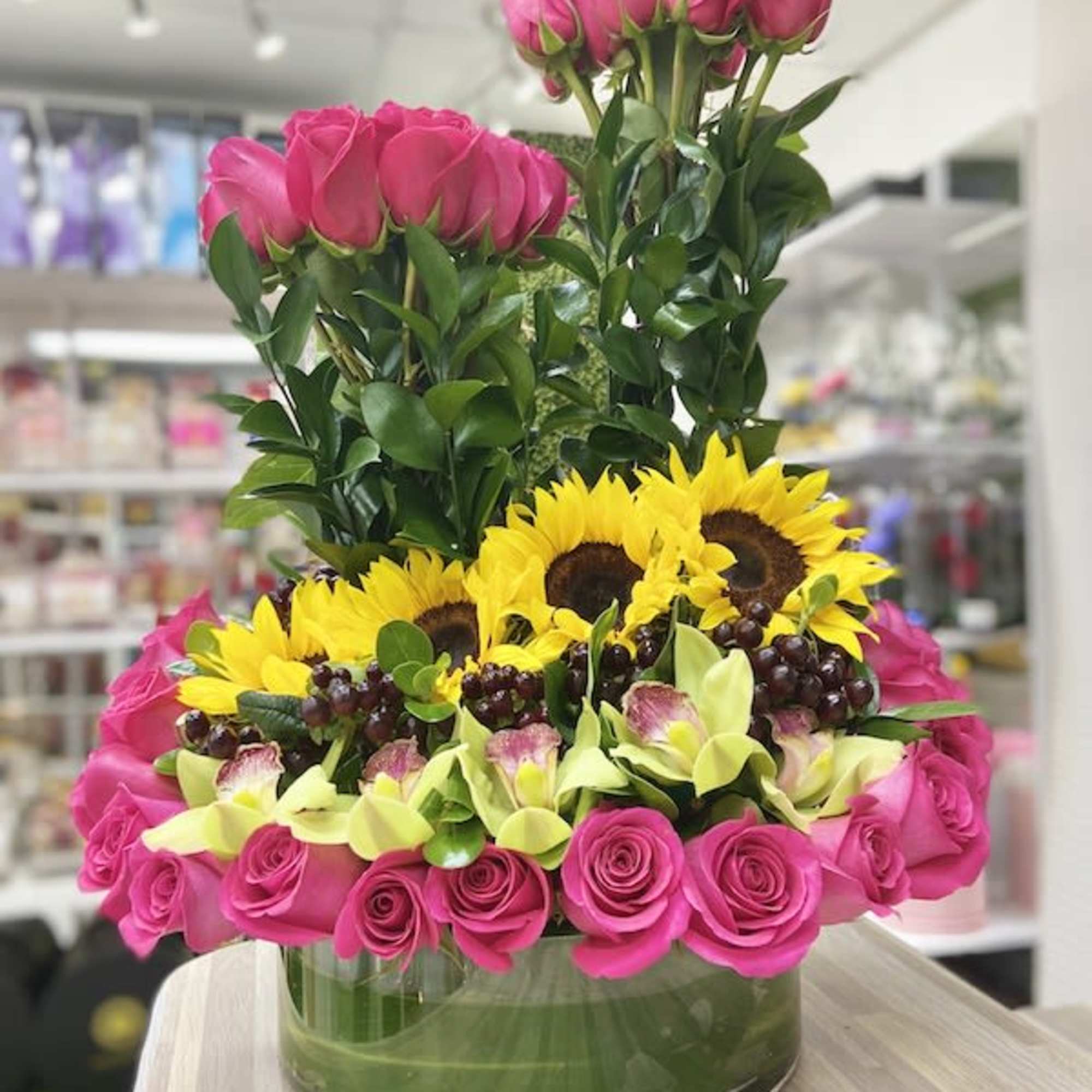 Low glass vase with hot pink roses, yellow sunflowers, green calla lilies, and grapes in a layered arrangement