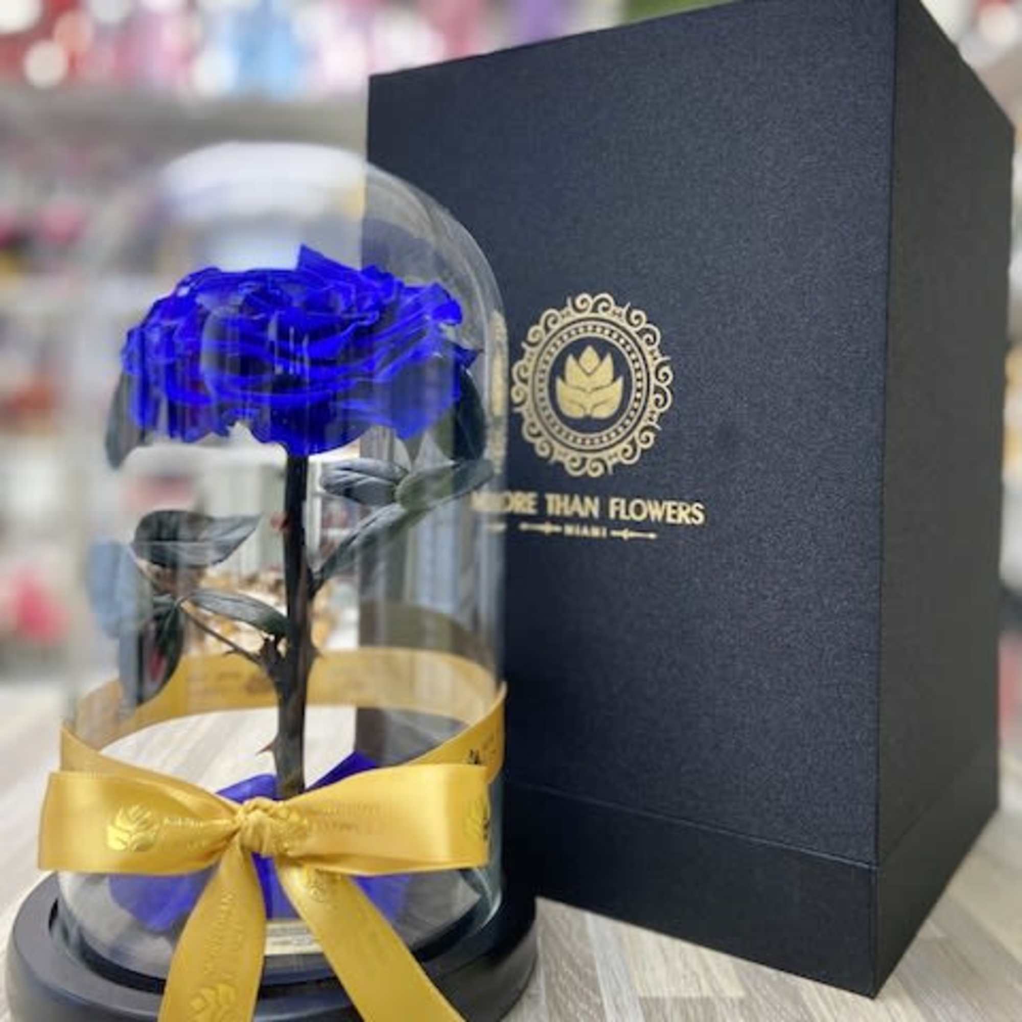 Single blue rose under a glass dome with yellow ribbon beside a tall black gift box.