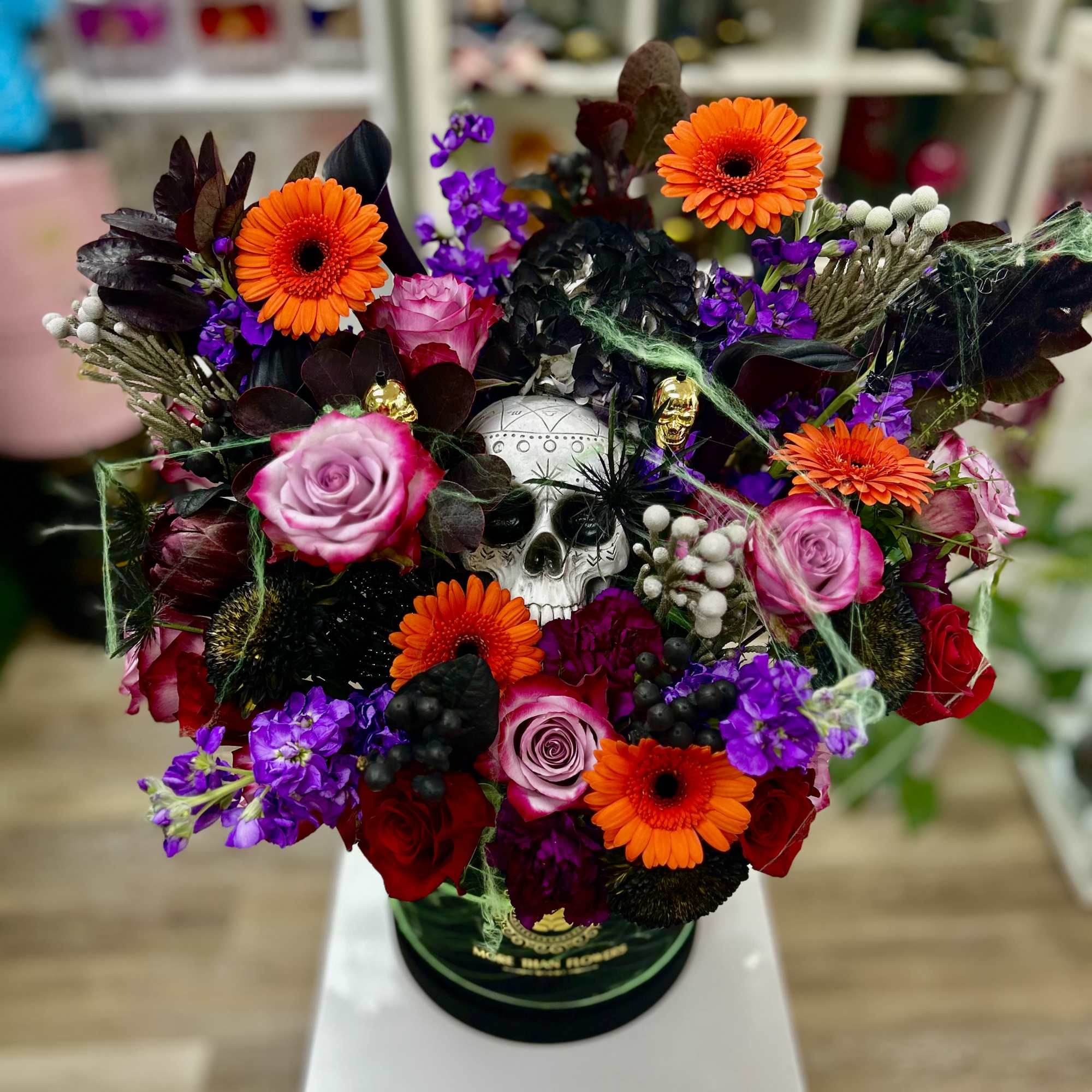 Vibrant orange, purple, and pink floral arrangement with a decorative skull and cobwebs in a green hatbox container