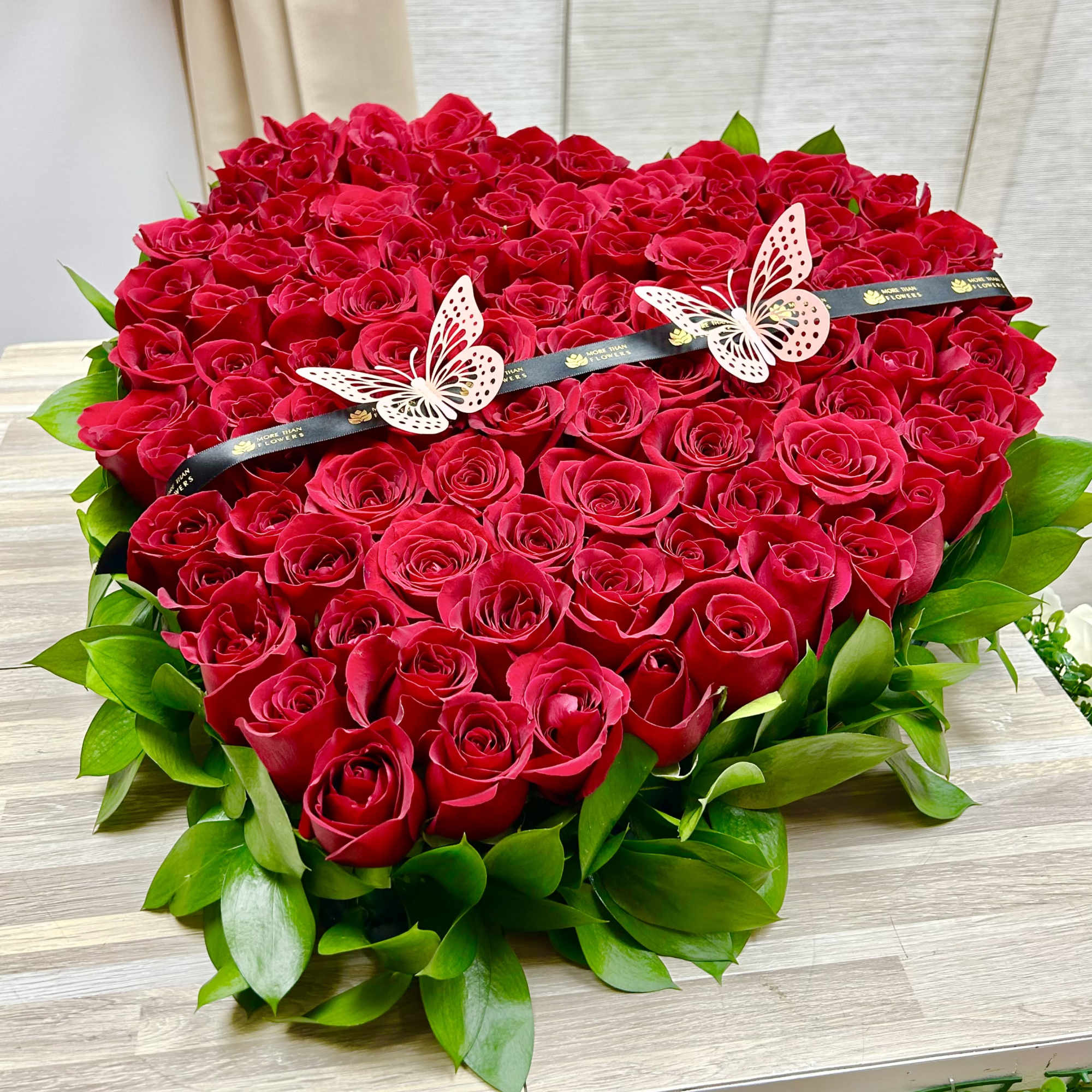 Heart-shaped arrangement of red roses framed with green foliage and decorated with butterflies.