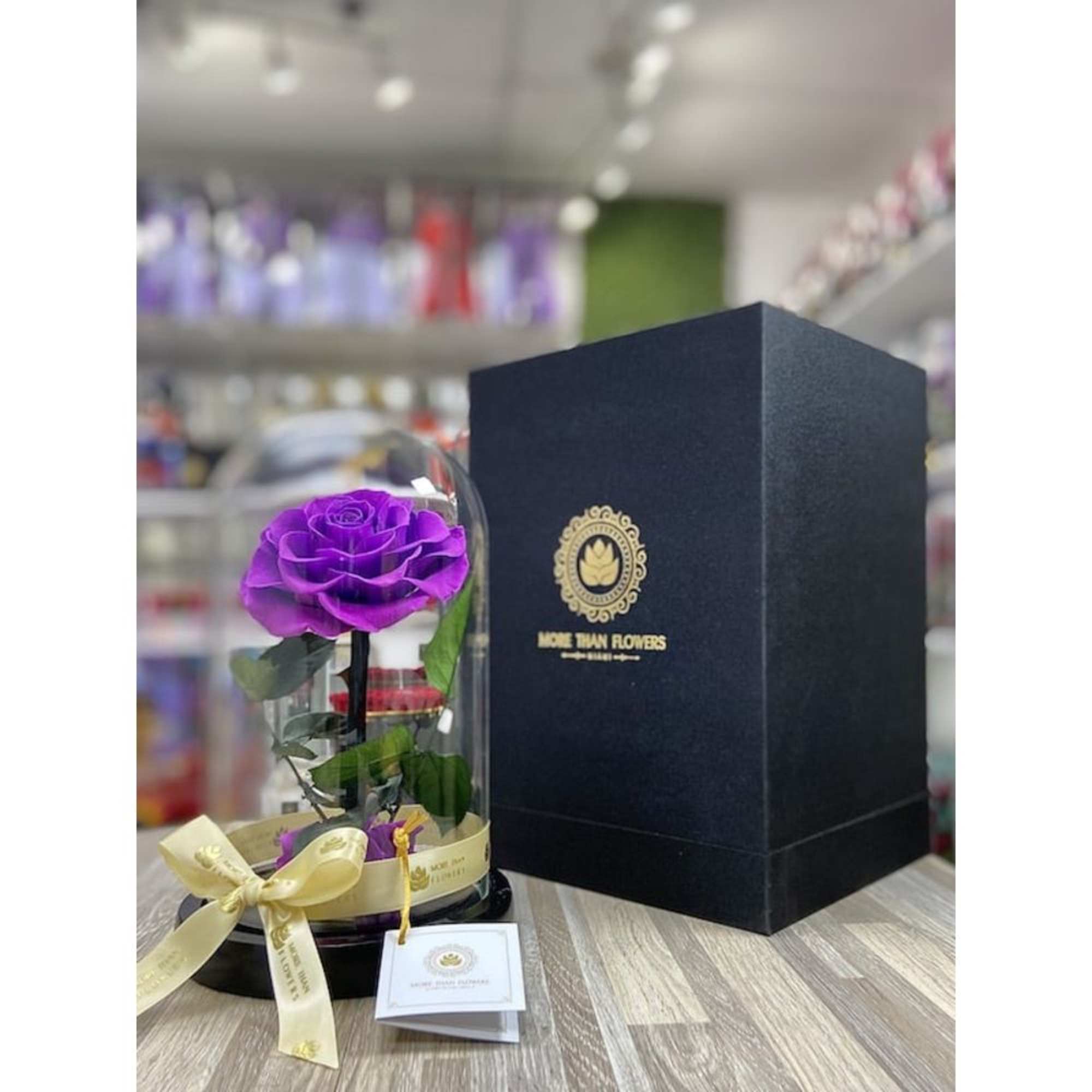 Single purple rose under a glass dome with ribbon on a black base beside a tall black gift box