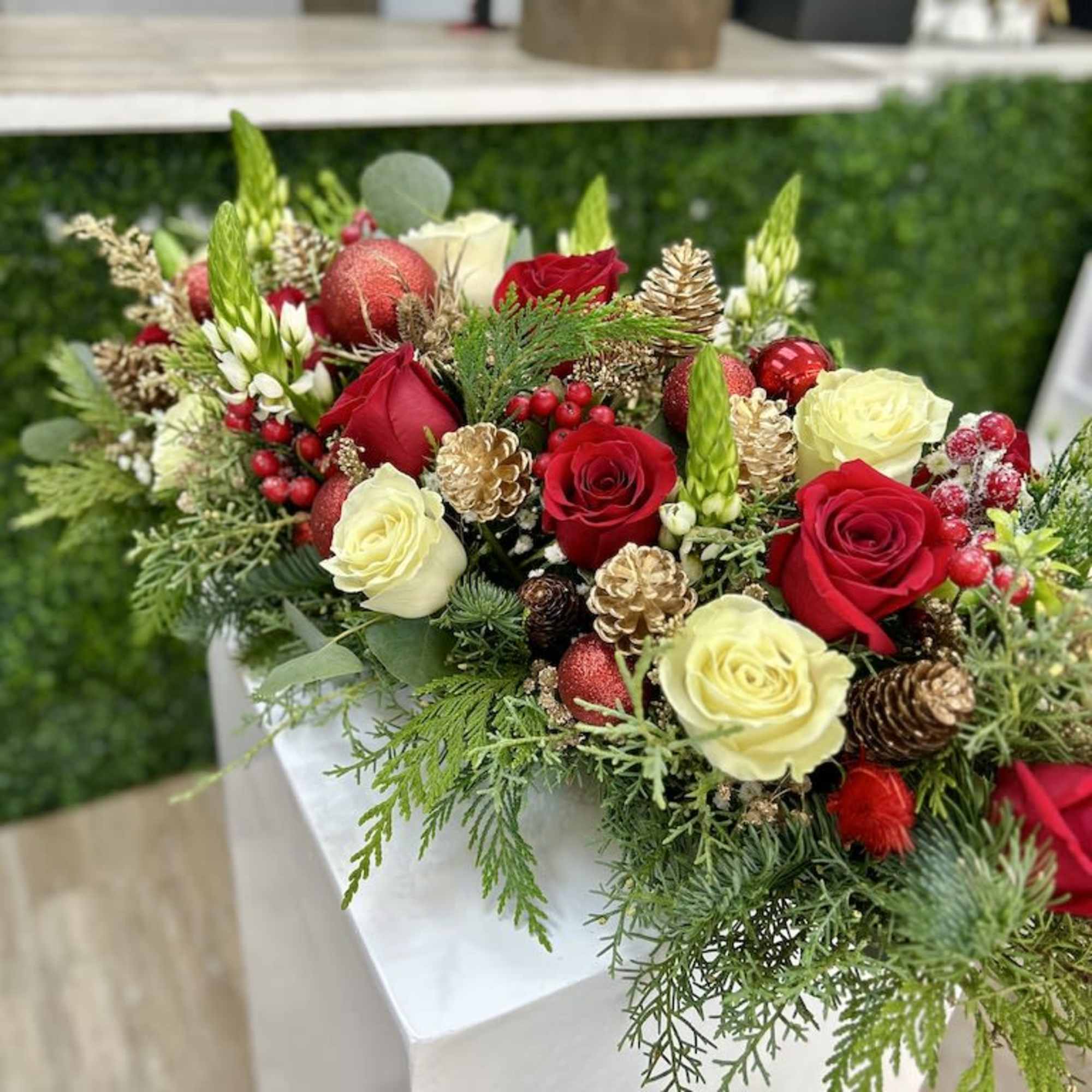 Long low arrangement of red and cream roses with evergreens, berries, and gold pine cones in a holiday style.