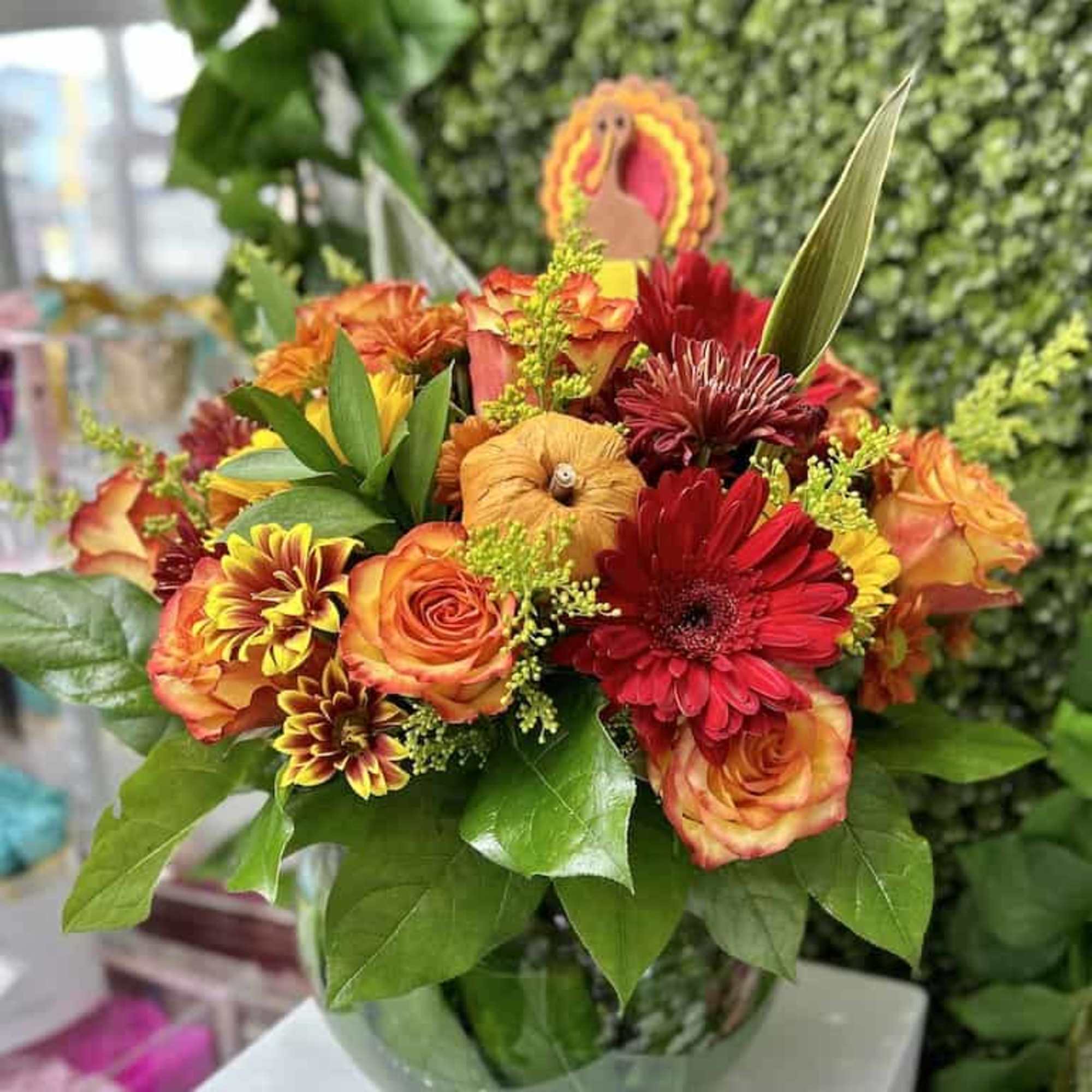 Fall arrangement of orange roses and red gerbera daisies in a round glass vase with a turkey pick