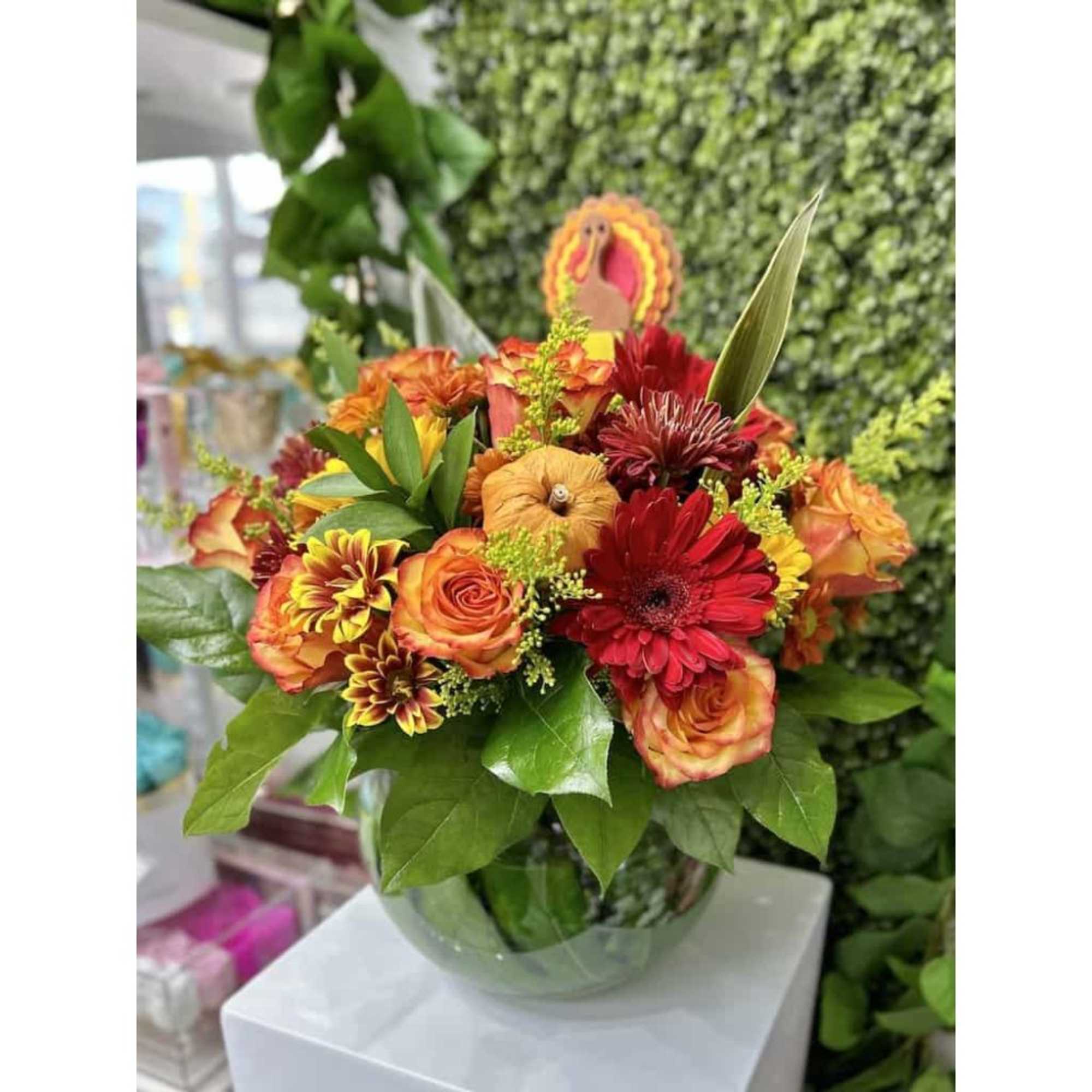 Low round arrangement of orange roses, red gerbera daisies, and yellow mums in a clear bowl vase with small pumpkins