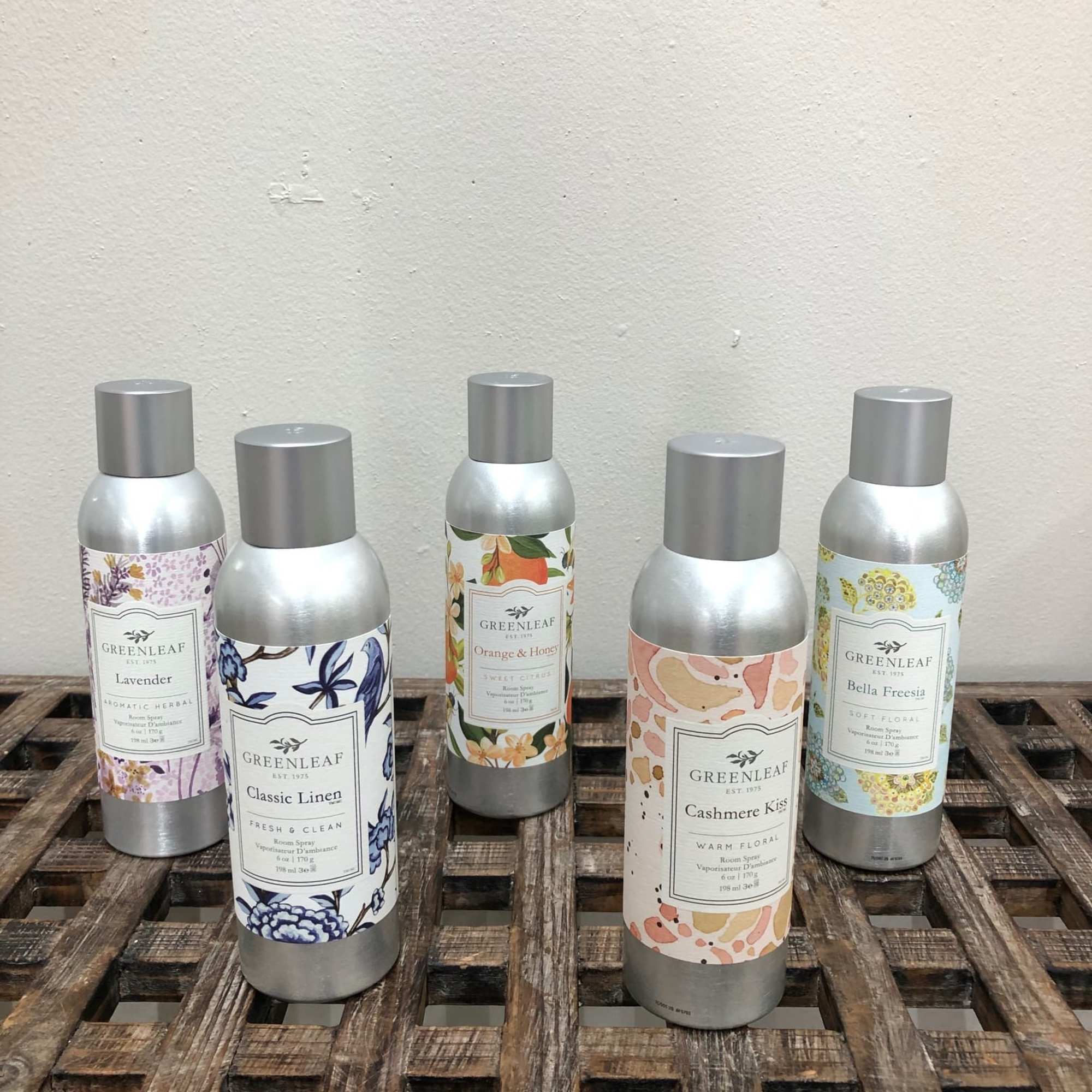 Set of five decorative scented spray cans on a wooden grid shelf against a light wall