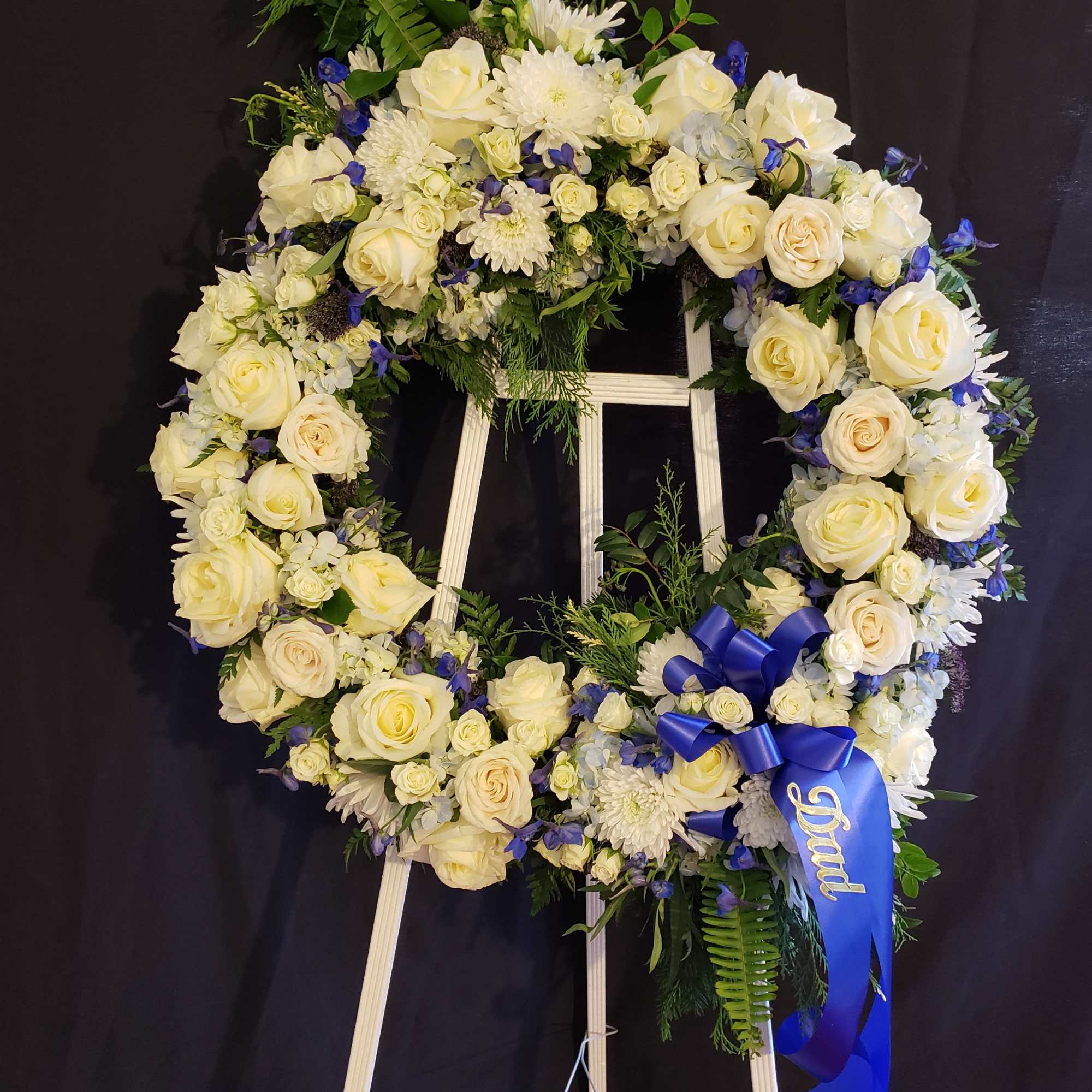 White rose and chrysanthemum funeral wreath on an easel with a blue ribbon