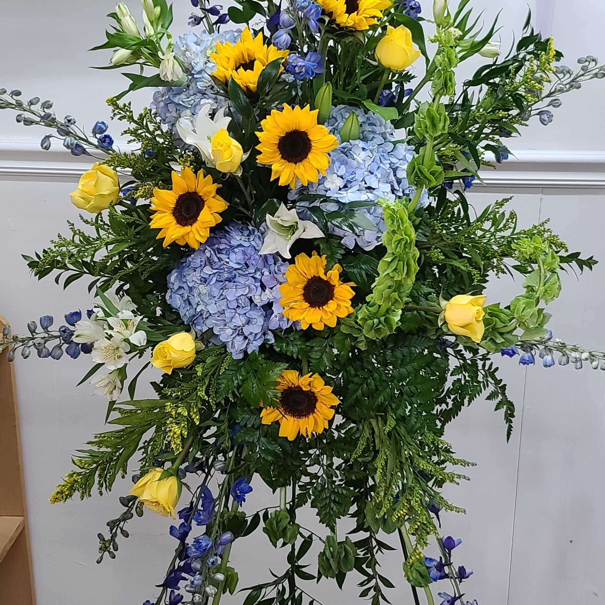 Traditional Standing spray of golden sunflowers, yellow roses, blue delphinium, bells of