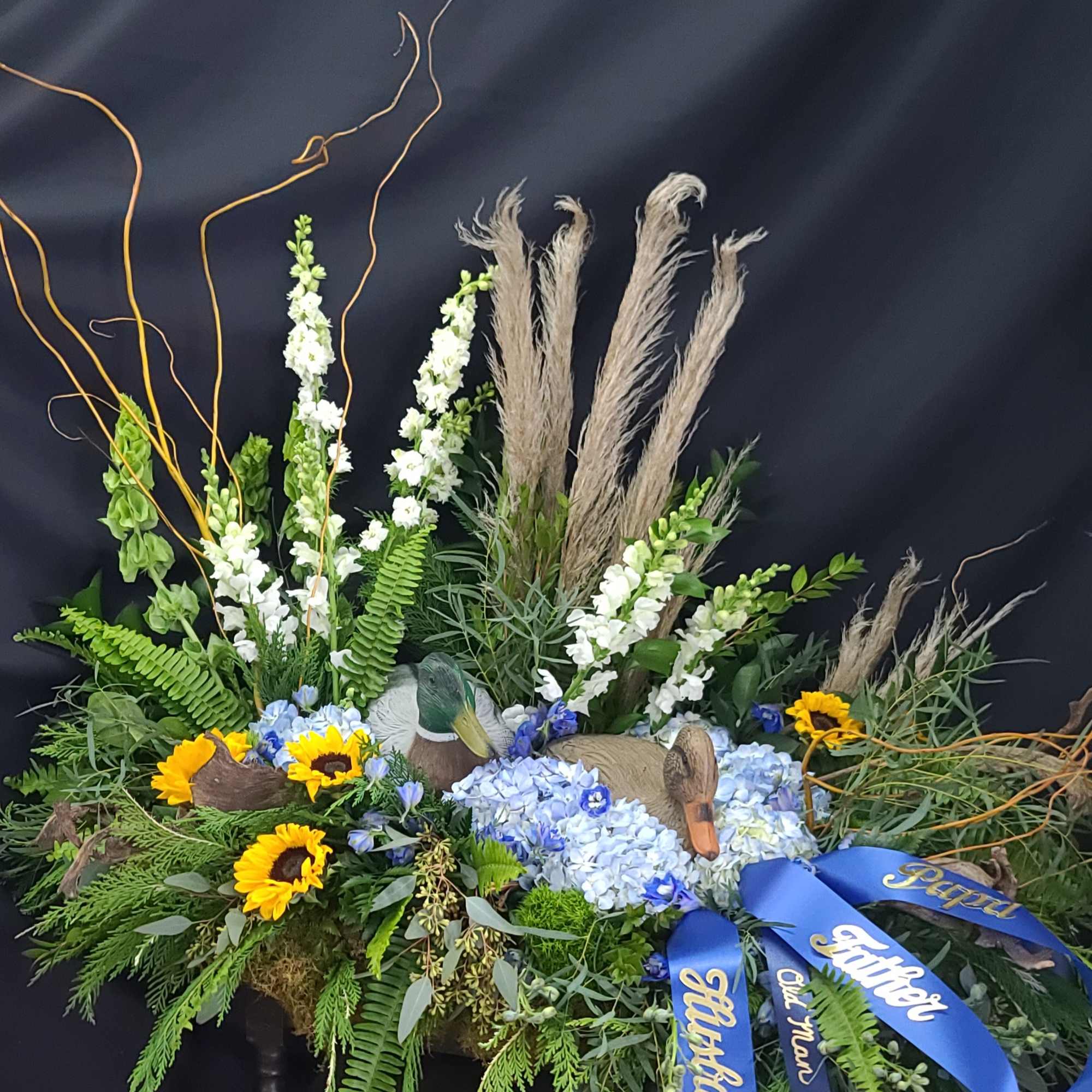 Floral tribute with sunflowers, blue hydrangeas, and blue ribbons on a dark stand