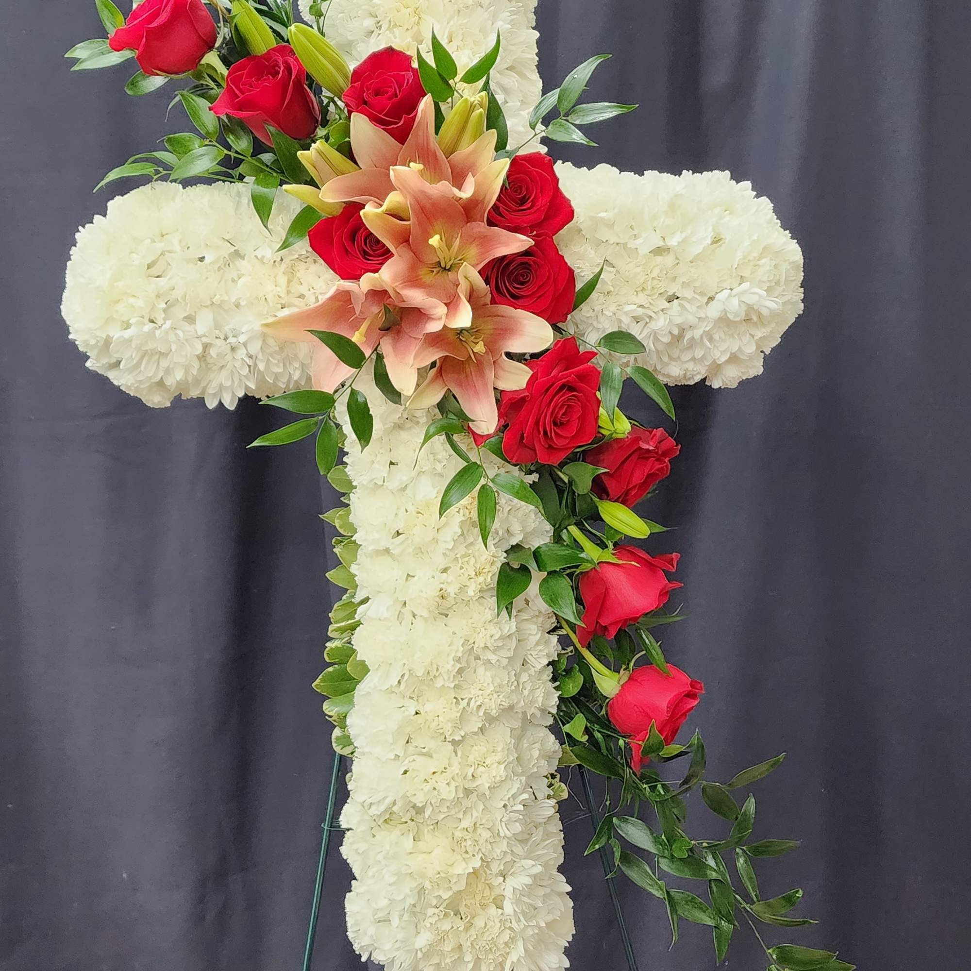White floral cross on a stand with red roses and peach lilies