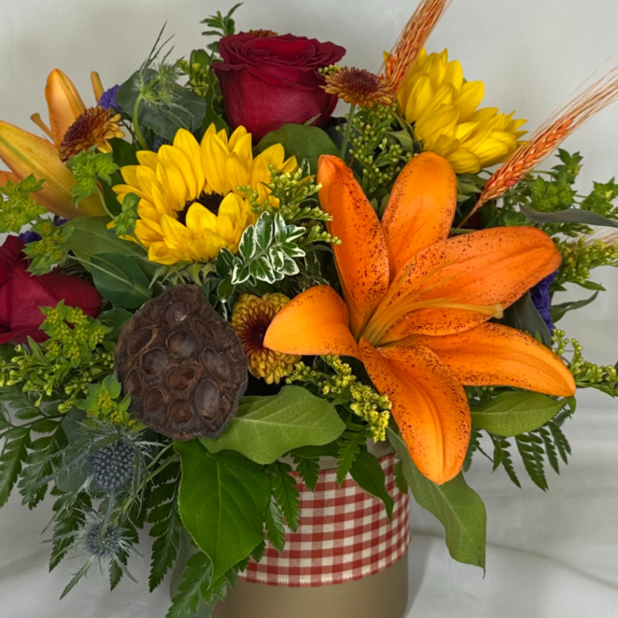 Mixed bouquet with orange lilies, red roses, and yellow sunflowers in a gingham-wrapped container.