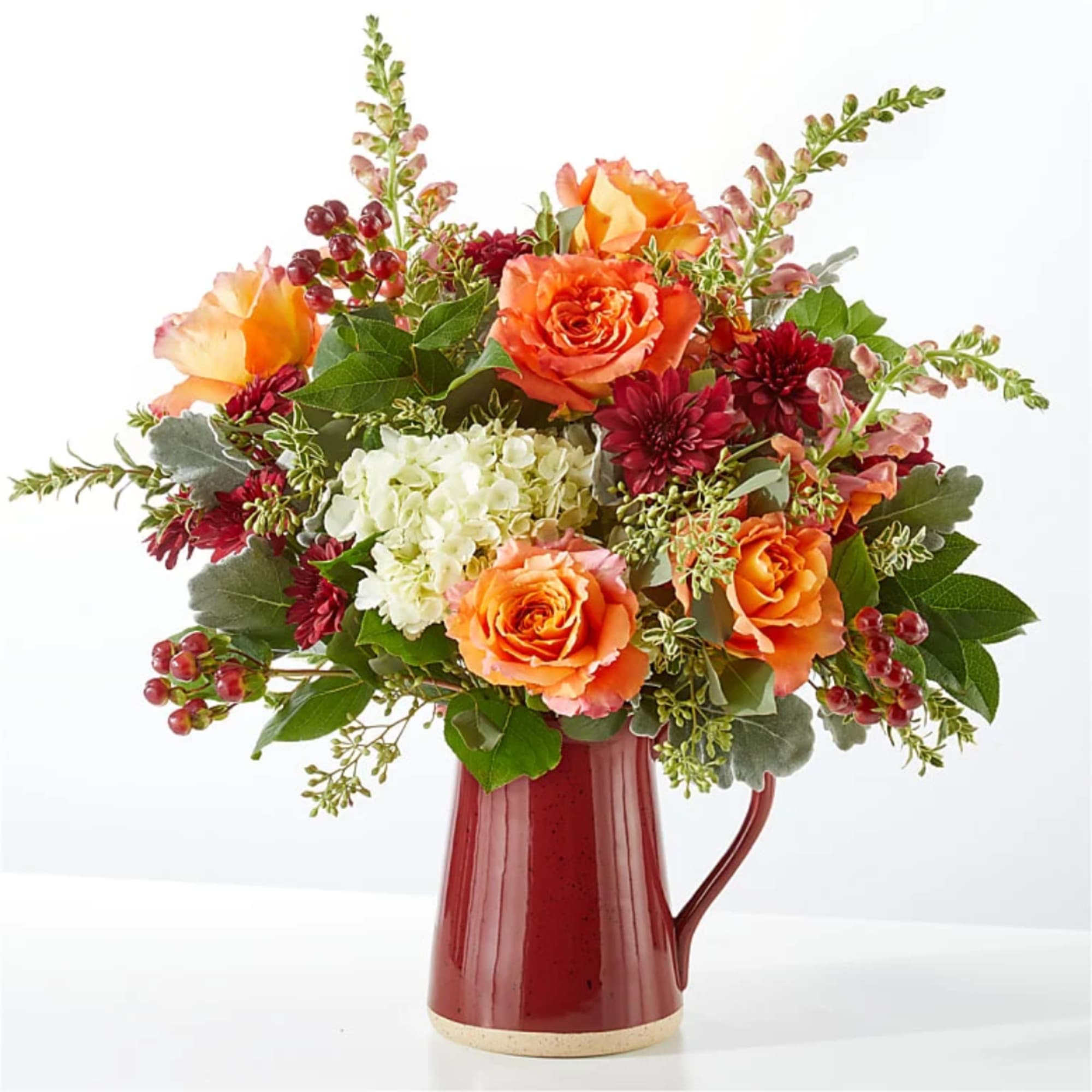 Orange roses and burgundy flowers arranged in a red pitcher vase