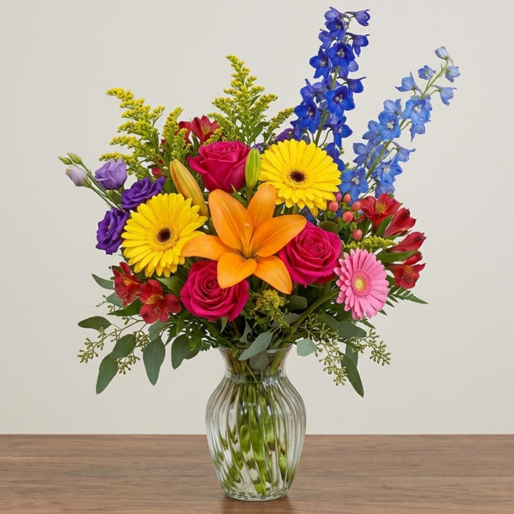 Mixed bouquet in a clear glass vase with roses, lilies, gerberas, and blue delphinium