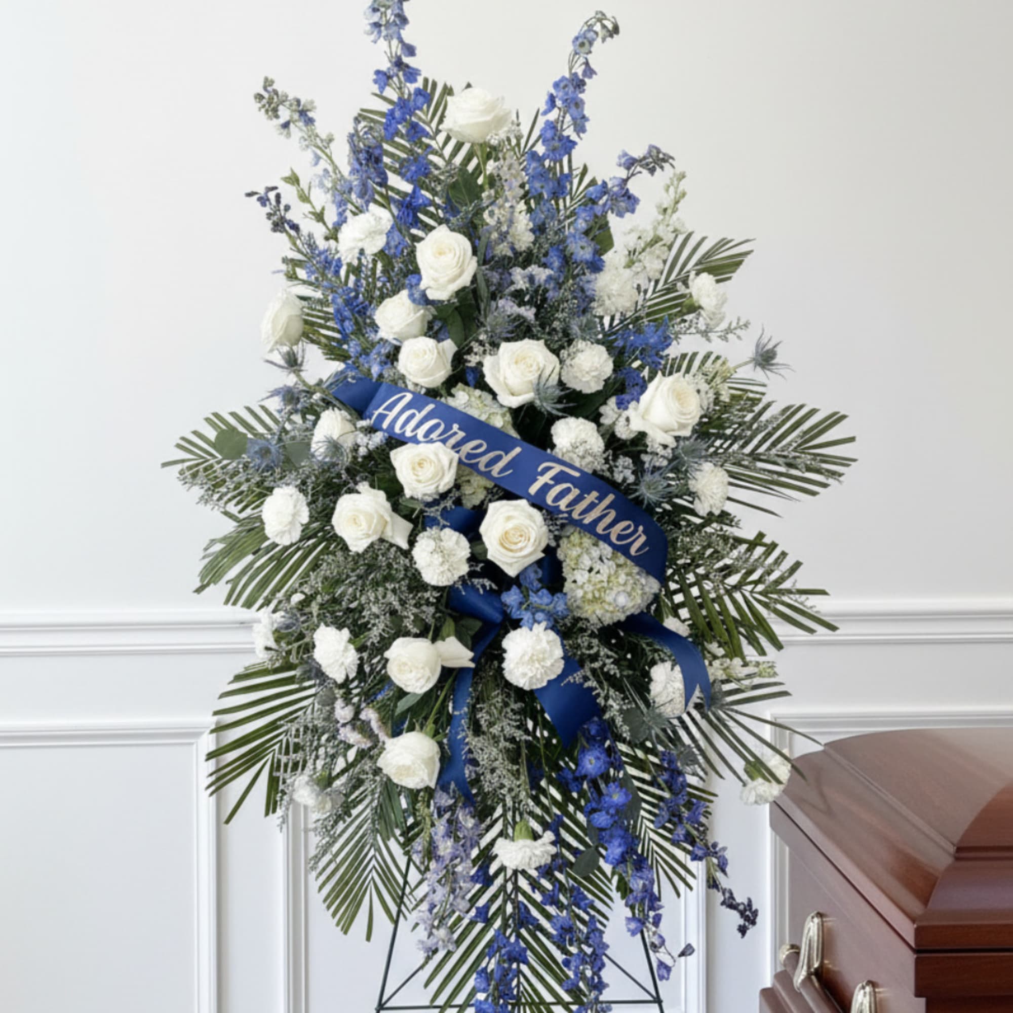 Tall white and blue funeral spray with a ribbon reading Adored Father