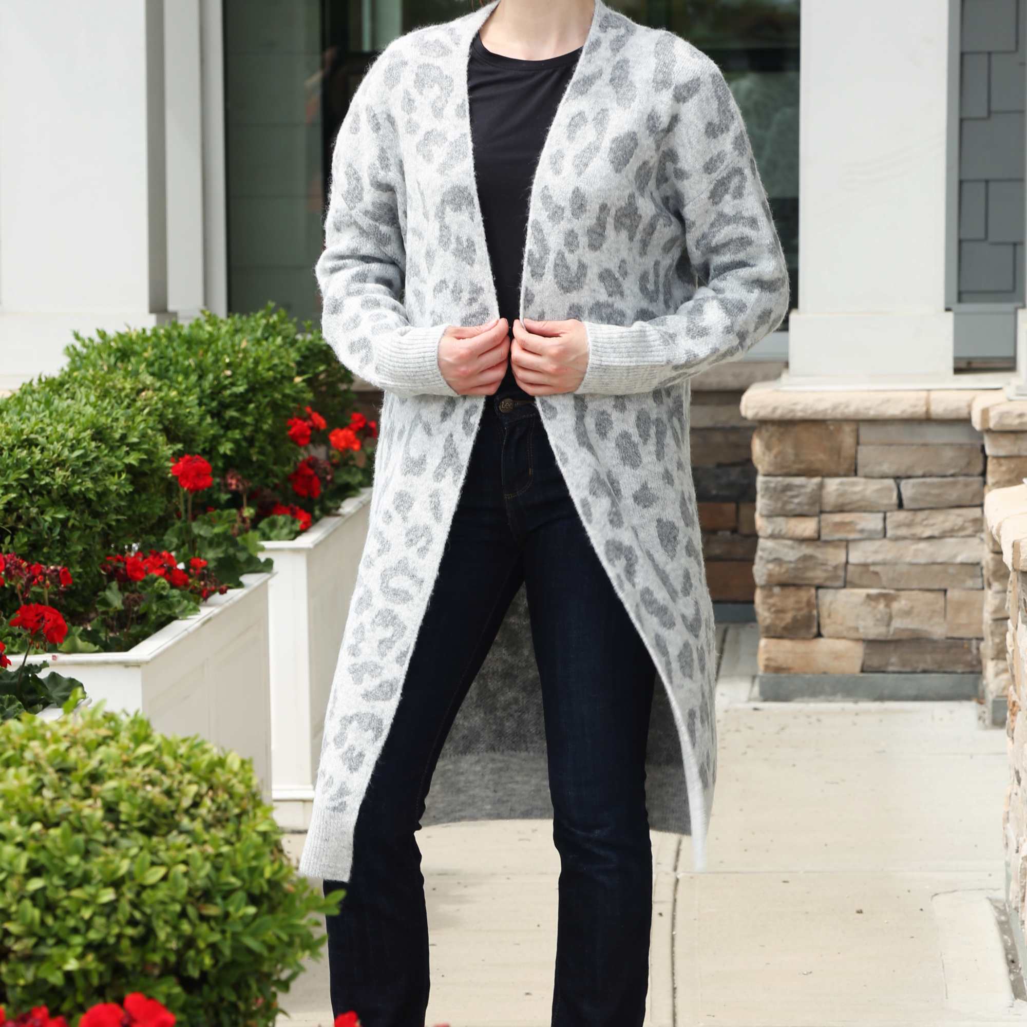 Woman in a long gray patterned cardigan and dark jeans standing by white planters with red flowers
